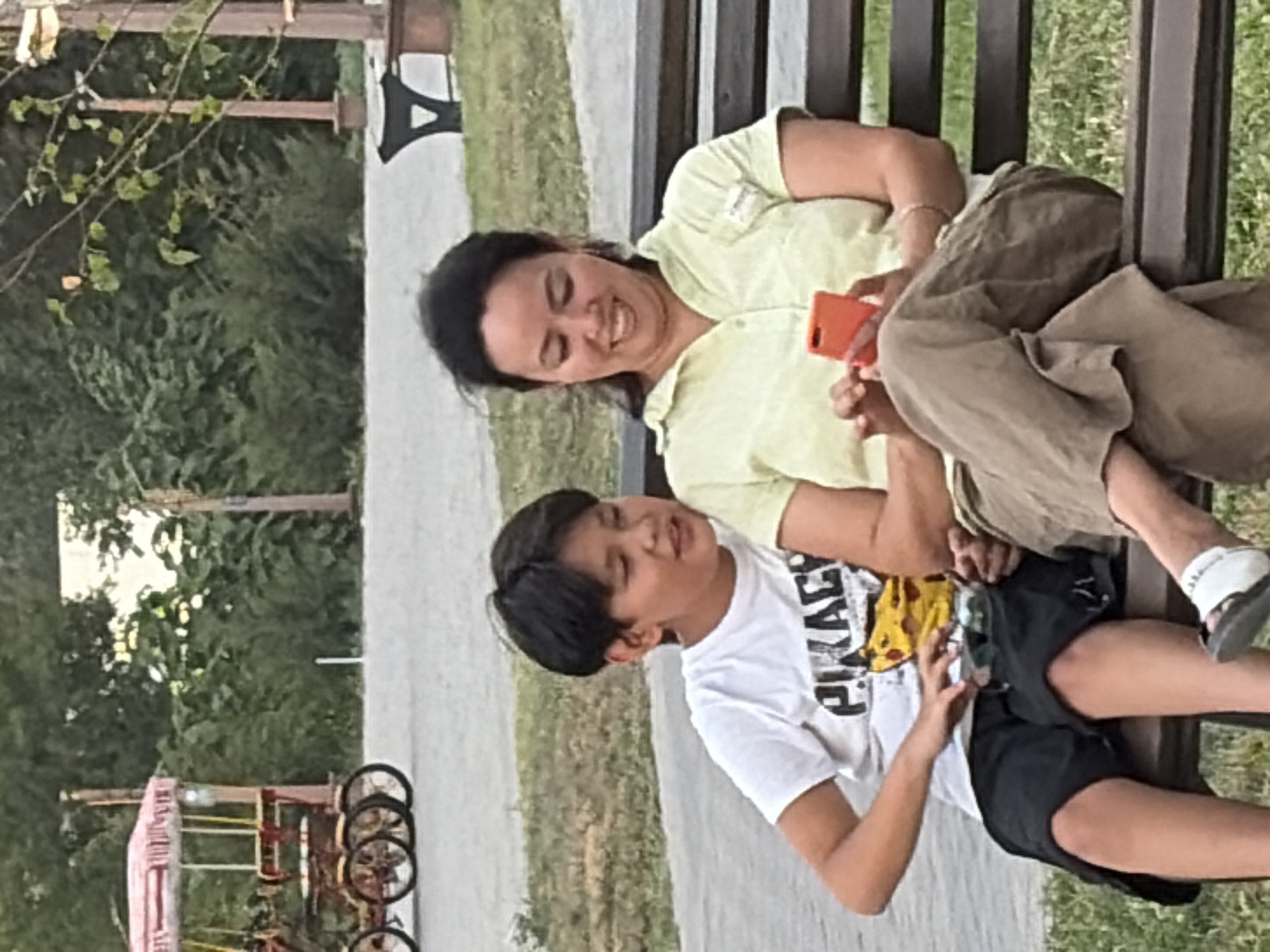 In an outdoor park setting, a young boy and a woman are seated side-by-side on a dark wooden bench. The boy, on the left, wears a white t-shirt with a cartoon character and the word "PIRACY" printed on it, along with black shorts. He is looking towards the woman. The woman, on the right, is wearing a light green short-sleeved collared shirt and brown pants. She is holding an orange smartphone in her hands and looking down at it, smiling. The background shows green foliage, trees, and a pathway. In the distance, there are elements of what appears to be a recreational park, possibly including a decorative carriage with wheels and canopy.