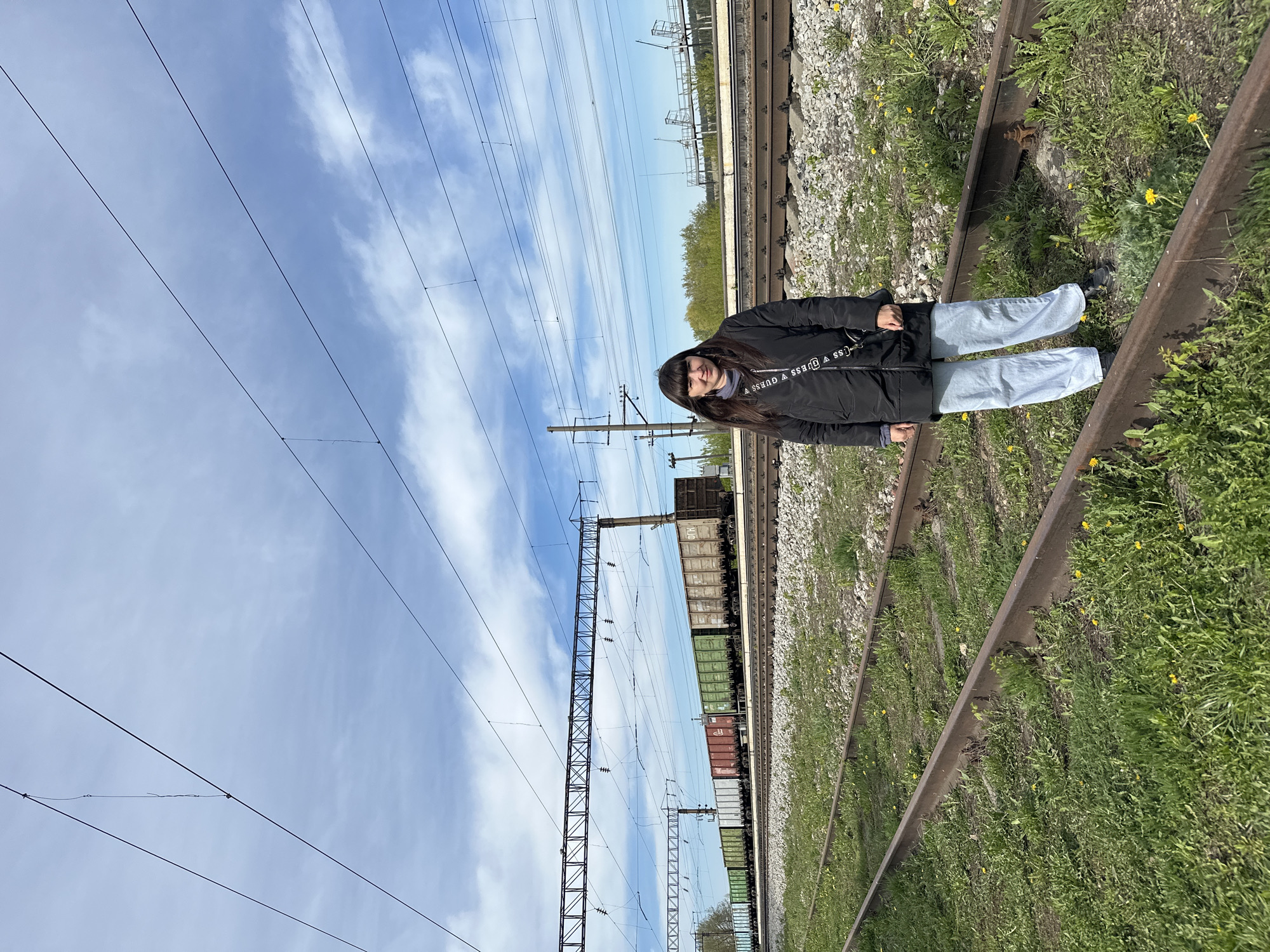 A medium shot captures a person standing on a railroad track. The individual, likely female, has long dark hair and is wearing a black jacket with repeating "GUESS" logos on the strap of a shoulder bag. Light-colored jeans and dark shoes complete the outfit.
The railroad track is overgrown with grass and weeds, interspersed with small yellow flowers. The adjacent gravel ballast supports the rails. In the background, several freight cars are visible on the track. Power lines are strung overhead, supported by metal towers. The sky is light blue with streaks of white clouds.
The image was taken outdoors on a sunny day near the city of Кечёво, Russia.