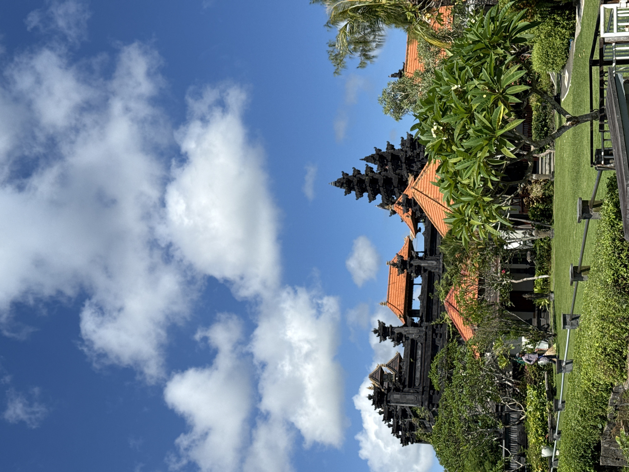 This image captures a vibrant outdoor scene, likely a traditional Balinese temple complex or resort, under a clear blue sky with scattered white cumulus clouds. The setting is characterized by lush tropical vegetation and distinctive architecture, consistent with Kuta Selatan, Indonesia.
The dominant subjects are several traditional Balinese structures featuring tiered black roofs, reminiscent of meru towers or temple gates, and buildings with warm orange-tiled roofs. These structures are interspersed throughout the mid-ground and background, partially framed by dense greenery. Large, well-maintained green lawns extend across the scene, bordered by hedges and various trees, including prominent frangipani trees with small white flowers and tall palm trees.
In the mid-ground, near a dark wooden fence or railing, two individuals are visible. They appear to be engaged in light activity, possibly maintenance or general work within the grounds. One person wears a light blue top and dark pants, while the other is dressed in a light-colored top and darker pants.
The bright sunlight indicates it is daytime, casting clear shadows and illuminating the vivid colors of the landscape. The overall atmosphere is one of tranquility and natural beauty, characteristic of a tropical paradise with rich cultural elements. No visible text is present in the image.
