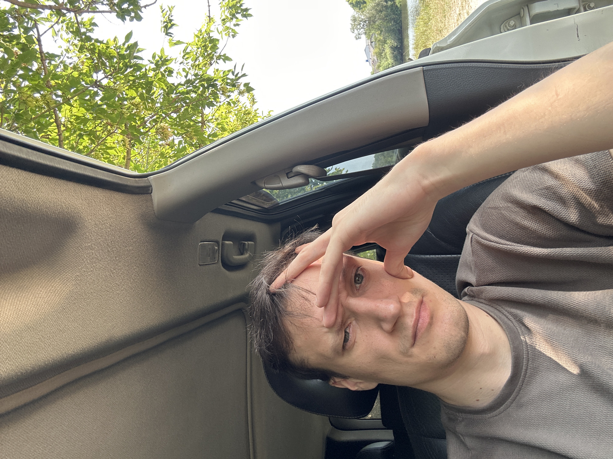 A man is sitting in the driver's seat of a car, looking directly at the camera with his right hand on his forehead and his left hand resting on his cheek. He is wearing a brown t-shirt. Sunlight filters through the green leaves of a tree outside the open car door, casting shadows on his face and shirt. In the background, a body of water and trees are visible under a bright sky. The car interior is visible, including the ceiling and part of the dashboard. The image appears to have been taken in a rural or natural setting, likely near a body of water.