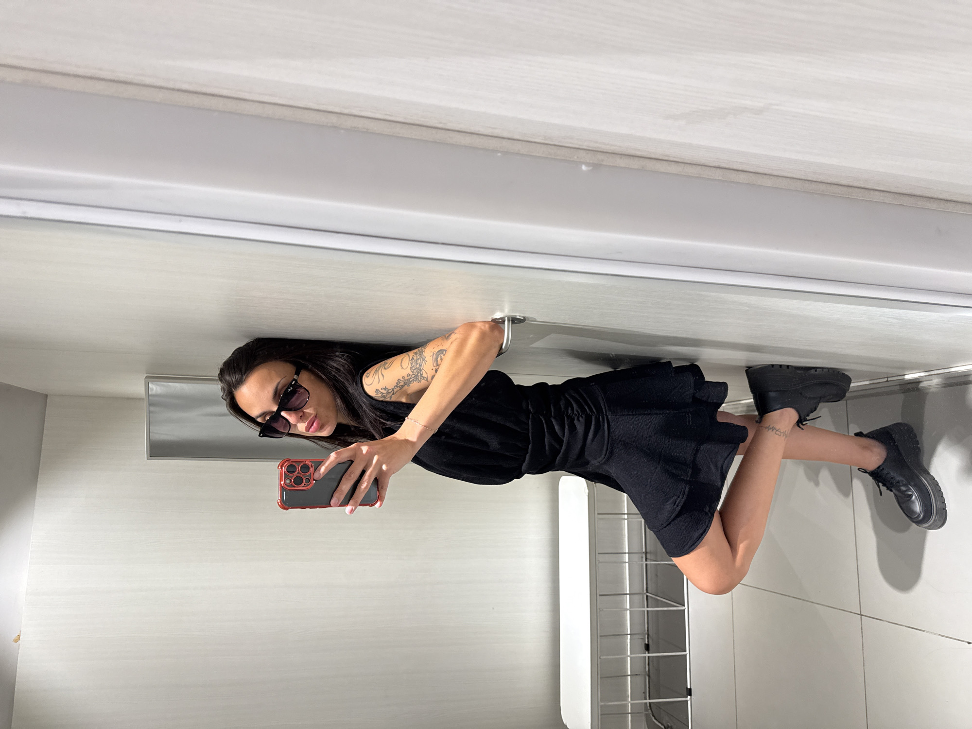 A young woman with dark hair and a tattoo on her left arm is taking a selfie in what appears to be a modern, minimalist interior. She is wearing a black dress and black combat boots. She is holding a smartphone with a red case. The background includes white walls, a white ceiling, a towel warmer, and light fixtures. There is no information available that confirms the location is in Pleven, Bulgaria.