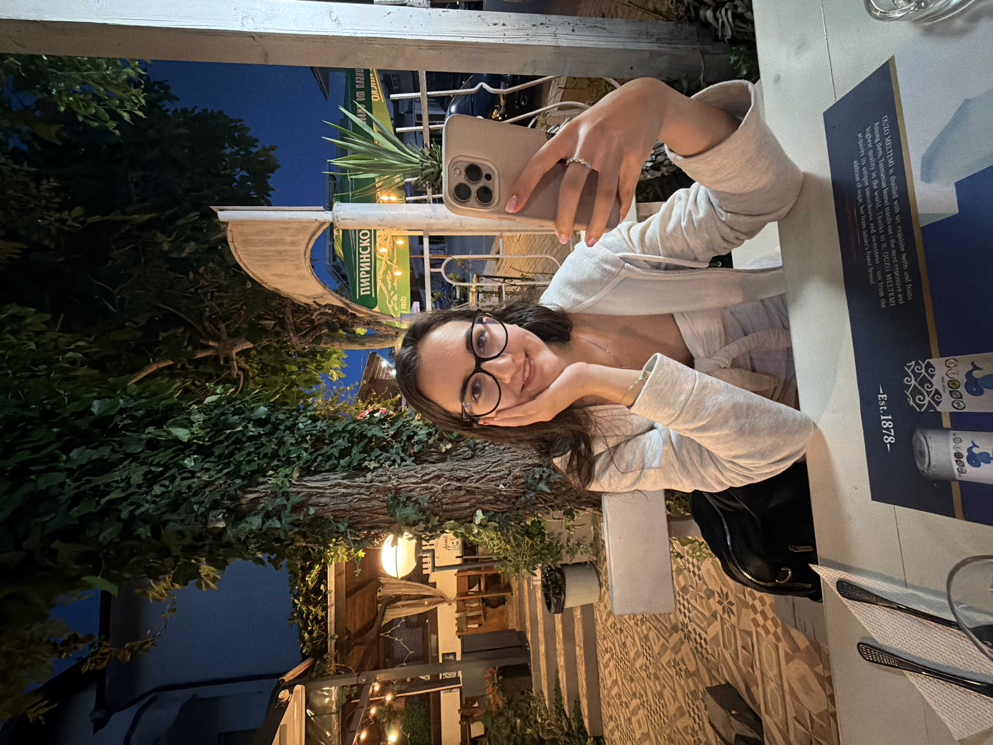 A young woman with dark hair and glasses is taking a selfie with her phone in an outdoor restaurant in Kiten, Bulgaria. She is wearing a light-colored hoodie and a white top. She is sitting at a white table, and in the background, there are trees, lights, and other restaurant seating. The scene appears to be at dusk or early evening.