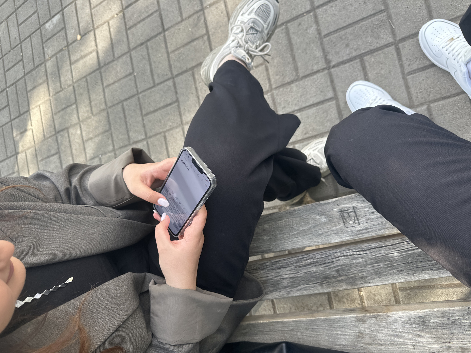 An eye-level shot shows two people sitting on a wooden bench with metal supports, with the view angled downwards towards their laps and feet.
The person on the left is wearing a gray blazer over black pants. The person's hands are holding a smartphone, and the screen displays an active messaging app. A silver necklace is partially visible below the blazer. The person on the right is wearing black pants.
Both individuals are wearing athletic shoes. The person on the left has on gray sneakers, and the person on the right is wearing white sneakers. The bench is placed on a paved area. The city of Yerevan, Armenia is not directly verifiable from the image.
