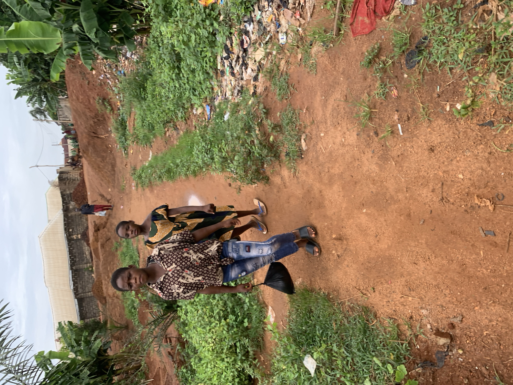 Two women are walking on a dirt path. The woman in front is wearing a patterned shirt and jeans and carrying a black object. The second woman is wearing a patterned dress. The path is bordered by vegetation and garbage. There is a building and a group of people visible further up the path. The location is Ukwu-Abwa, Nigeria.
