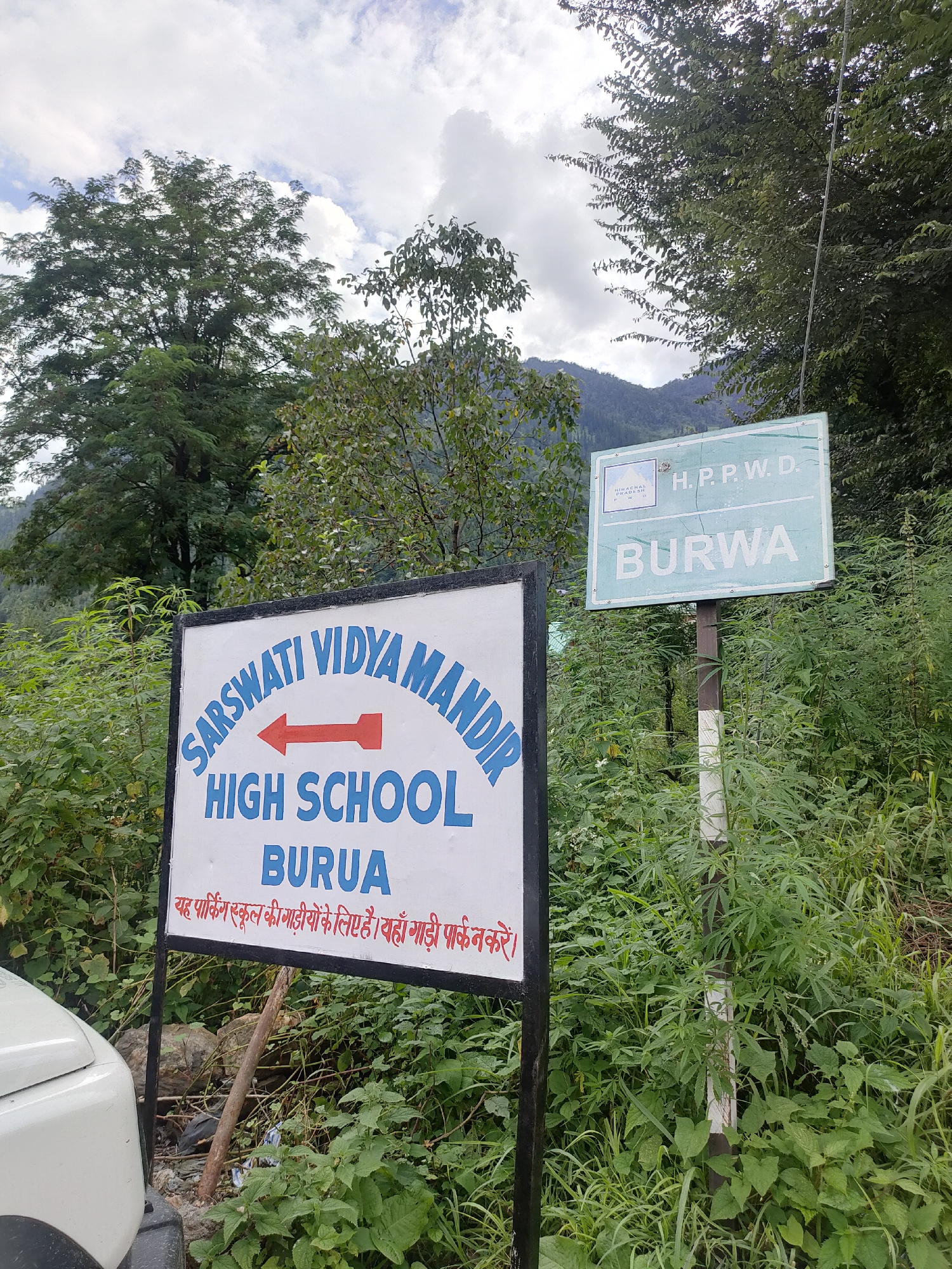 This is an outdoor, eye-level, medium shot of two signs in Burwa, India. The sign on the left is white with a black border and displays "SARSWATI VIDYA MANDIR HIGH SCHOOL BURUA" in blue text. A red arrow points left above "HIGH SCHOOL." Below "BURUA," there is text in Hindi stating, "This parking is for school vehicles. Park your vehicle there." The sign on the right is a pale green rectangle with a white border and black text. It reads "H.P.P.W.D." at the top and "BURWA" in large letters below. A small crest with "HIMACHAL PRADESH PWD" is in the upper left corner of this sign. The signs are surrounded by lush green foliage and trees. A white vehicle is partially visible on the left edge of the frame. The sky is cloudy.