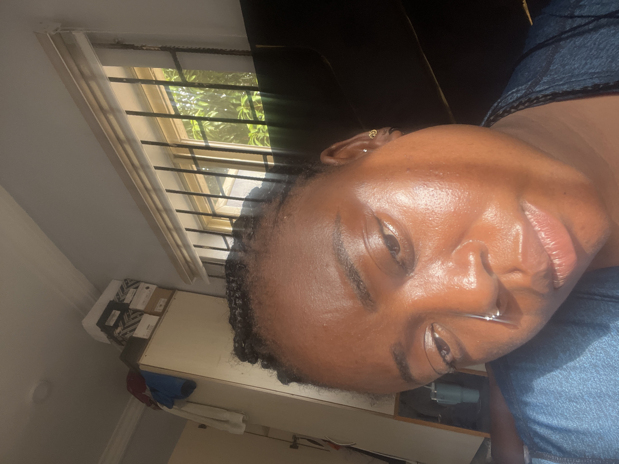 The image features a close-up portrait of a person, likely female, with dark skin and short, dark, braided hair visible at the hairline. She has a metallic septum nose piercing and small gold hoop earrings in both ears. Her face is well-lit, displaying highlights on her skin, suggesting it is either oily, sweaty, or has a product applied, and is illuminated by strong natural light. She wears a blue denim-like top or dress. Her expression is calm and relaxed, with a subtle, slight smile.
The setting appears to be an indoor room in Kubwa, Nigeria. In the background to the left, a window with horizontal metal security bars is visible, through which lush green foliage and bright sunlight can be seen, indicating a sunny daytime. Below the window, there is a light-colored piece of furniture, possibly a desk or shelf, on which several boxes and some folded clothes (blue and red) are placed. A part of a blue object, possibly an appliance, is also visible lower in the frame. The room has plain, light-colored walls. No visible text is present.