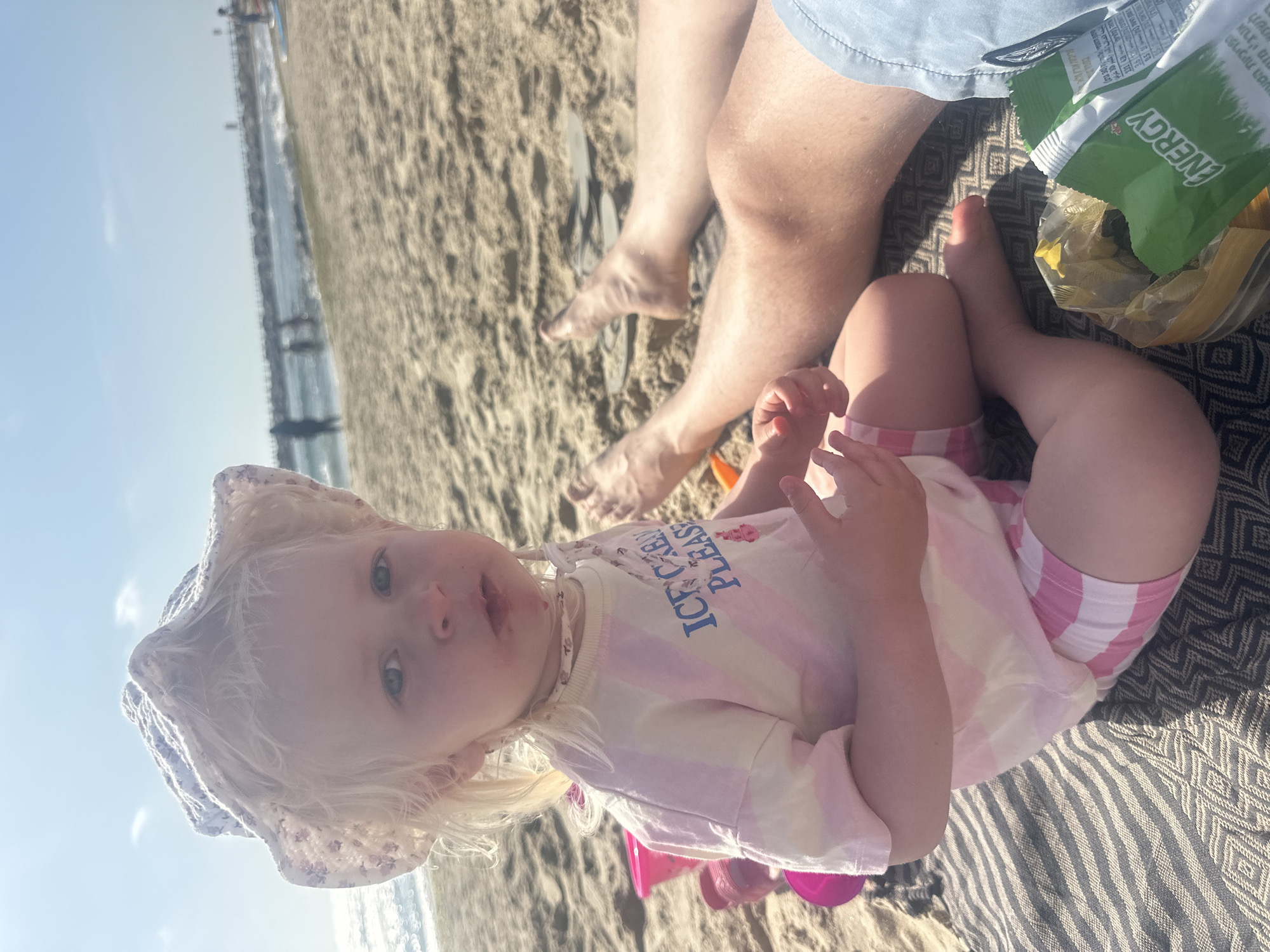 A young child, with fair skin and blonde hair, is sitting on a patterned blanket on a sandy beach. The child wears a floral bucket hat and a striped pink and white shirt that says "ICE CREAM PLEASE." The child is looking directly at the camera with a curious expression. Beside the child are the legs of an adult wearing light blue shorts, and a bag of chips with the word "ENERGY" visible on the packaging. The backdrop shows a beach scene, with sand, water, and possibly a distant structure in the water, and the clear sky suggests it is daytime. The location of the scene is on a beach in Tel Aviv, Israel.