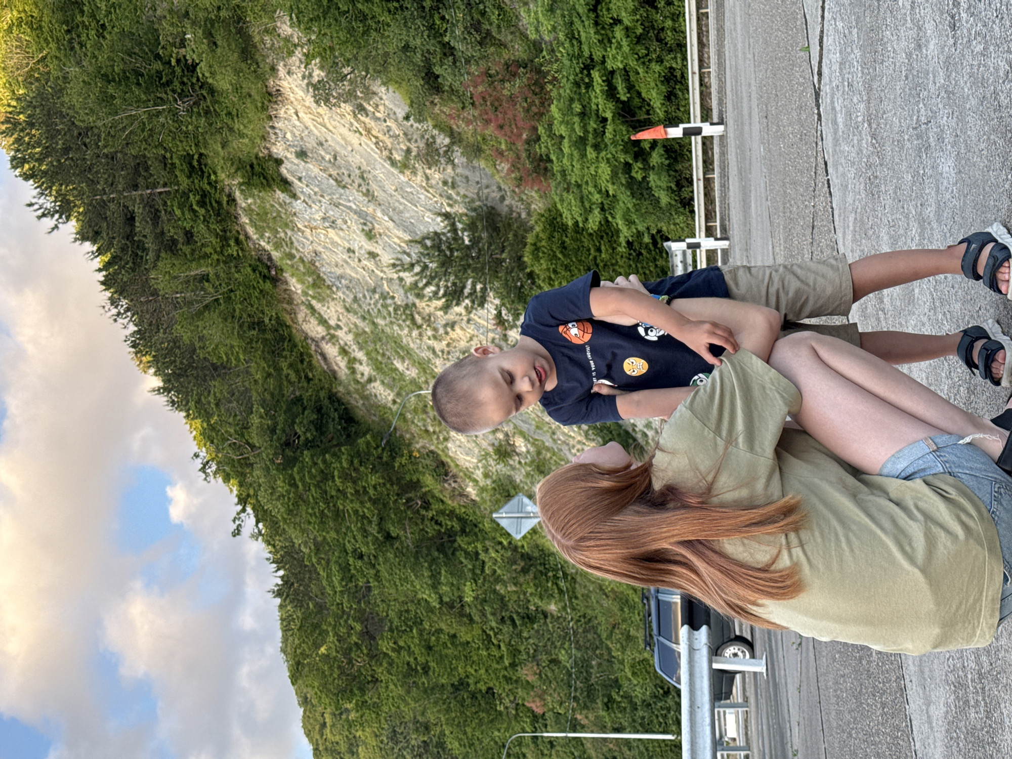 In Nebug, Russia, a young boy with short brown hair, wearing a dark blue t-shirt with cartoon graphics and light brown shorts, is interacting with a young woman with long red hair. The woman is crouched down, facing the boy, and appears to be wearing a light green t-shirt and denim shorts. They are standing on a paved surface, possibly a roadside or parking area, with a white metal guardrail to their left. In the background, a steep, tree-covered mountain slope rises, with a rocky embankment visible. A dark-colored SUV is parked on the left side of the frame. The sky above is partly cloudy.