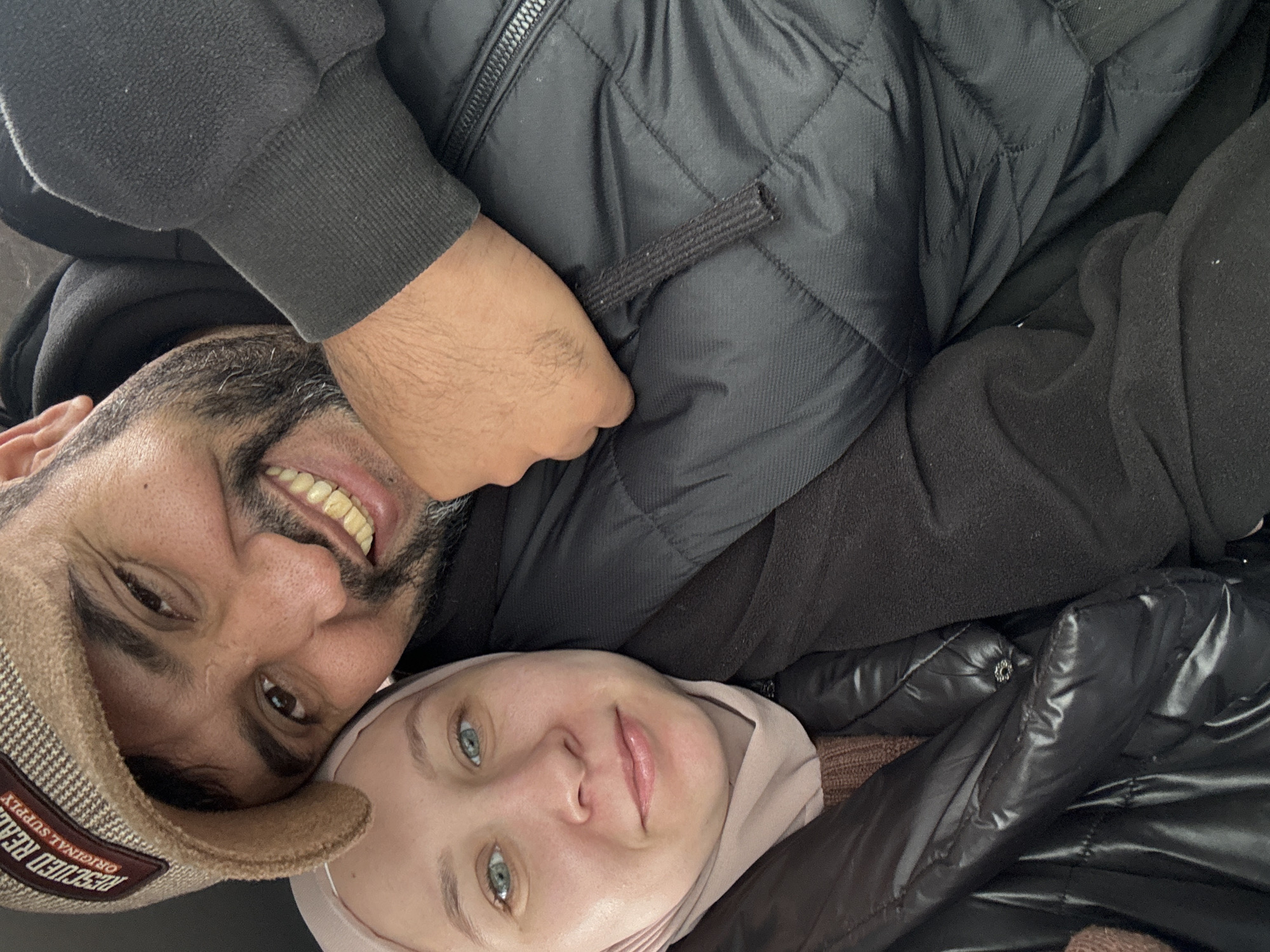 The image displays a close-up, rotated portrait of two individuals, likely captured as a selfie. The perspective suggests they are in close proximity, possibly lying down or leaning against each other.
On the left side of the frame (or top, in its current orientation), a man with dark hair and a beard is visible, smiling broadly and showing his teeth. He wears a light brown and dark-patterned cap with a brim that reads "RESCUED REAL ORIGINAL SUPPLY." He is also dressed in a dark, possibly black, hooded sweatshirt or jacket. His right arm, covered by a dark sleeve, is visible.
To the right (or bottom) of the man, a woman is seen with light blue-green eyes and a soft, serene expression. She wears a light pink or beige head covering, which appears to be a hijab, and a dark, shiny, quilted puffer jacket. A textured brown garment is visible beneath her jacket.
Both individuals are wearing warm, layered clothing, including heavy jackets and head coverings, which is suitable for cold weather. The atmosphere conveyed is intimate and content, with both subjects showing pleasant expressions. The location context for this scene is Chaikovsky, Russia, where such warm attire would be typical due to the climate. No specific time of day is indicated.