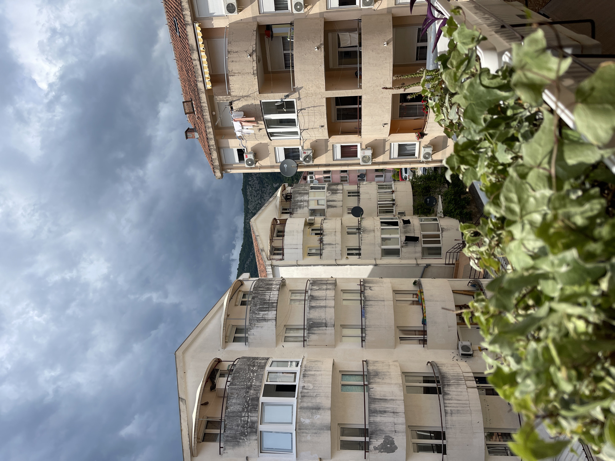 The image shows a view upwards between two apartment buildings in Budva, Montenegro. The building on the left is off-white and has rounded balconies. The building on the right is a light tan colour. Both buildings have satellite dishes and air conditioning units attached to their exteriors. Laundry is drying on a balcony. Dense green foliage is visible on the right. The sky is cloudy. A mountain is visible in the distance between the buildings.