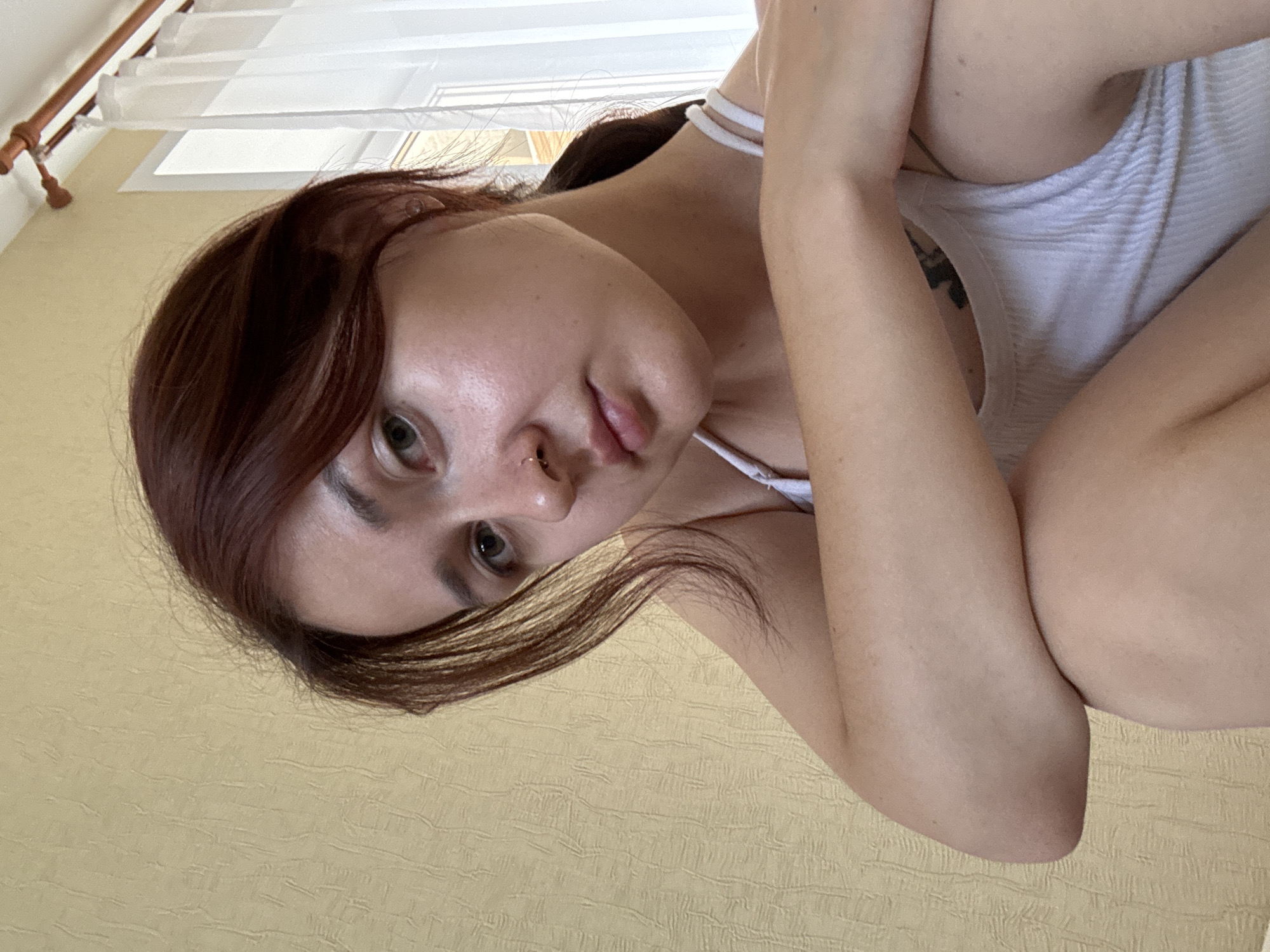 A young woman with auburn hair is pictured in a close-up, upper-body shot. She is wearing a white ribbed tank top and has a nose ring. Her arms are crossed in front of her chest. The background is a textured, pale yellow wall, with a white sheer curtain and a wooden curtain rod visible on the right side of the frame. The lighting suggests an indoor setting, possibly a bedroom or living area, with natural light coming from a window. The overall scene is casual and appears to be a selfie. The location is not specified in the image itself, and therefore cannot be determined to be in Naberezhnye Chelny, Russia.