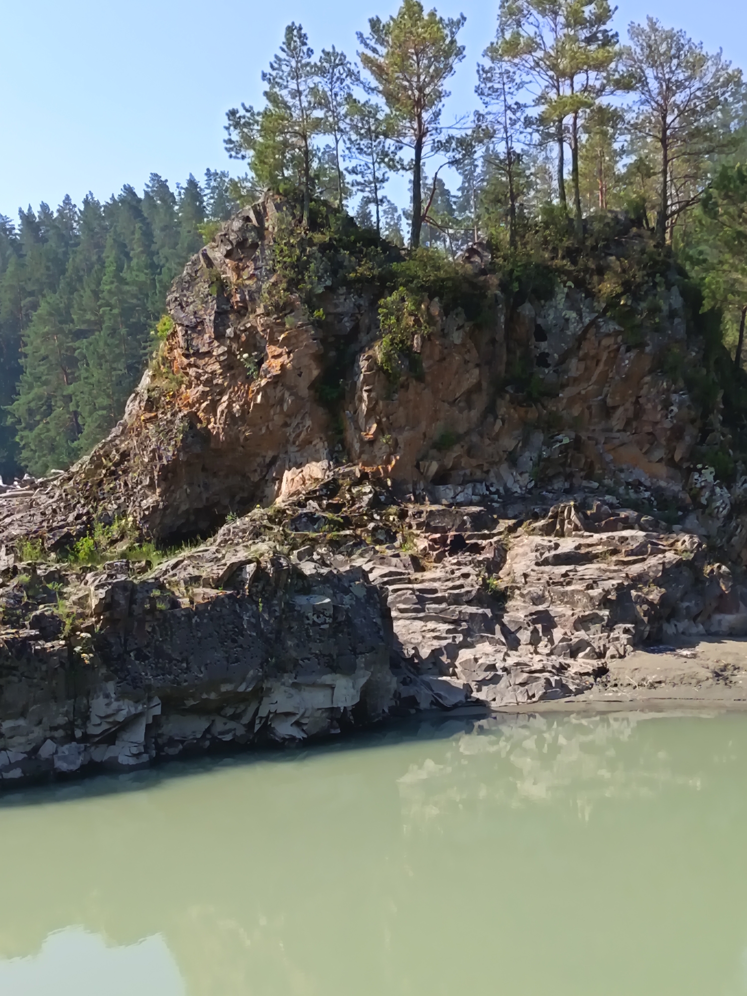 A photograph captures a rugged, rocky outcrop overlooking a pale green body of water. The outcrop is covered in sparse vegetation and several tall, slender pine trees with dark green needles. The rocks are layered and appear weathered, with some sections showing a reddish-brown hue. The water's surface is calm and reflects the sky and the rocks. In the background, a dense forest of pine trees stretches across the horizon under a clear blue sky. The image appears to be taken on a sunny day. The location is identified as Nizhnekayancha, Russia. There are no people or animals visible in the image.