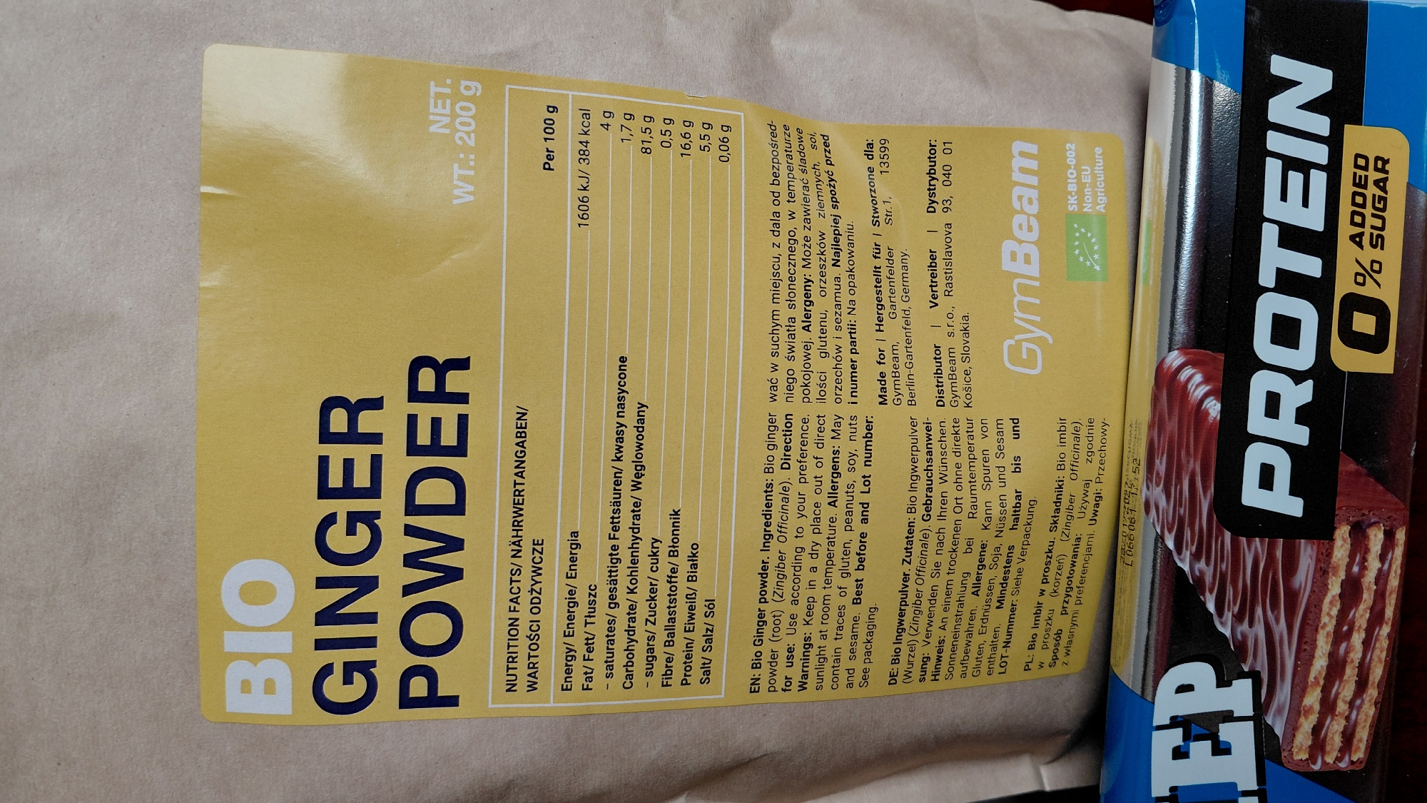A slightly rotated overhead shot features a partially visible package of "Bio Ginger Powder" (200g) and part of a "Protein 0% Added Sugar" bar. The ginger powder packaging, primarily yellow, lists nutritional information in multiple languages. The protein bar is blue, silver, and brown, with a close-up image of a chocolate-like waffle on the packaging.