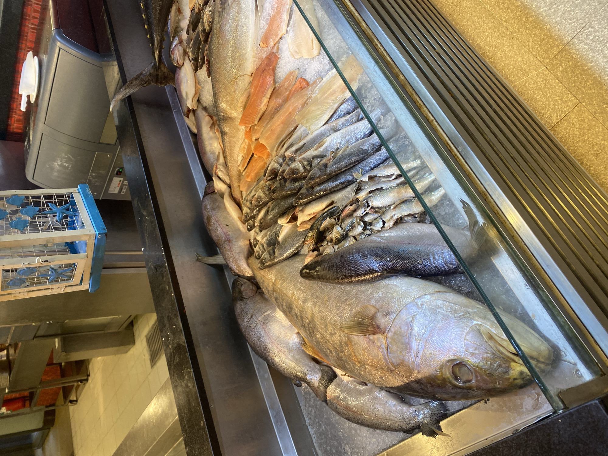 The image shows a close-up view of a display of fresh fish on ice at what appears to be a market or fishmonger. The fish are arranged in rows, with some whole fish and some filleted portions. The scale ranges from small, silvery fish to larger, more prominently featured fish. The setting is indoors, with a stainless steel counter and glass enclosure. In the background, there is a glimpse of shelving units and some equipment, suggesting a commercial food service environment. The image does not contain any specific visual information to pinpoint its location within Aksu, Turkey.