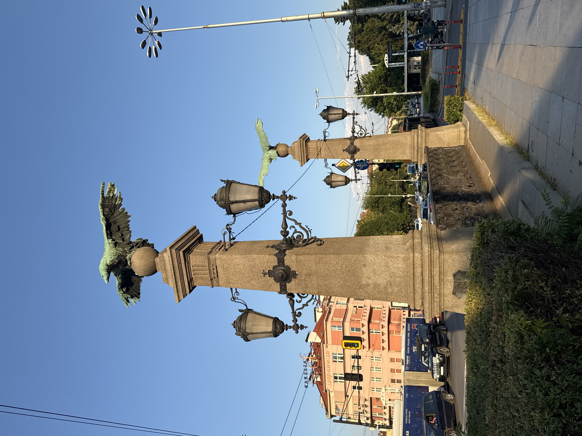 The image depicts the Eagles' Bridge in Sofia, Bulgaria, on a clear day. In the foreground, a large stone lamppost with ornate metalwork and four lamps is prominently featured. Atop the lamppost are two bronze eagle statues, one larger and positioned higher up, both with outstretched wings. To the left, a pink building with multiple windows and balconies is visible. The street is busy with traffic, including cars, a bus, and a motorcycle. Pedestrians are present on the sidewalk, and traffic lights are visible. The overall environment suggests an urban setting with historical architectural elements.