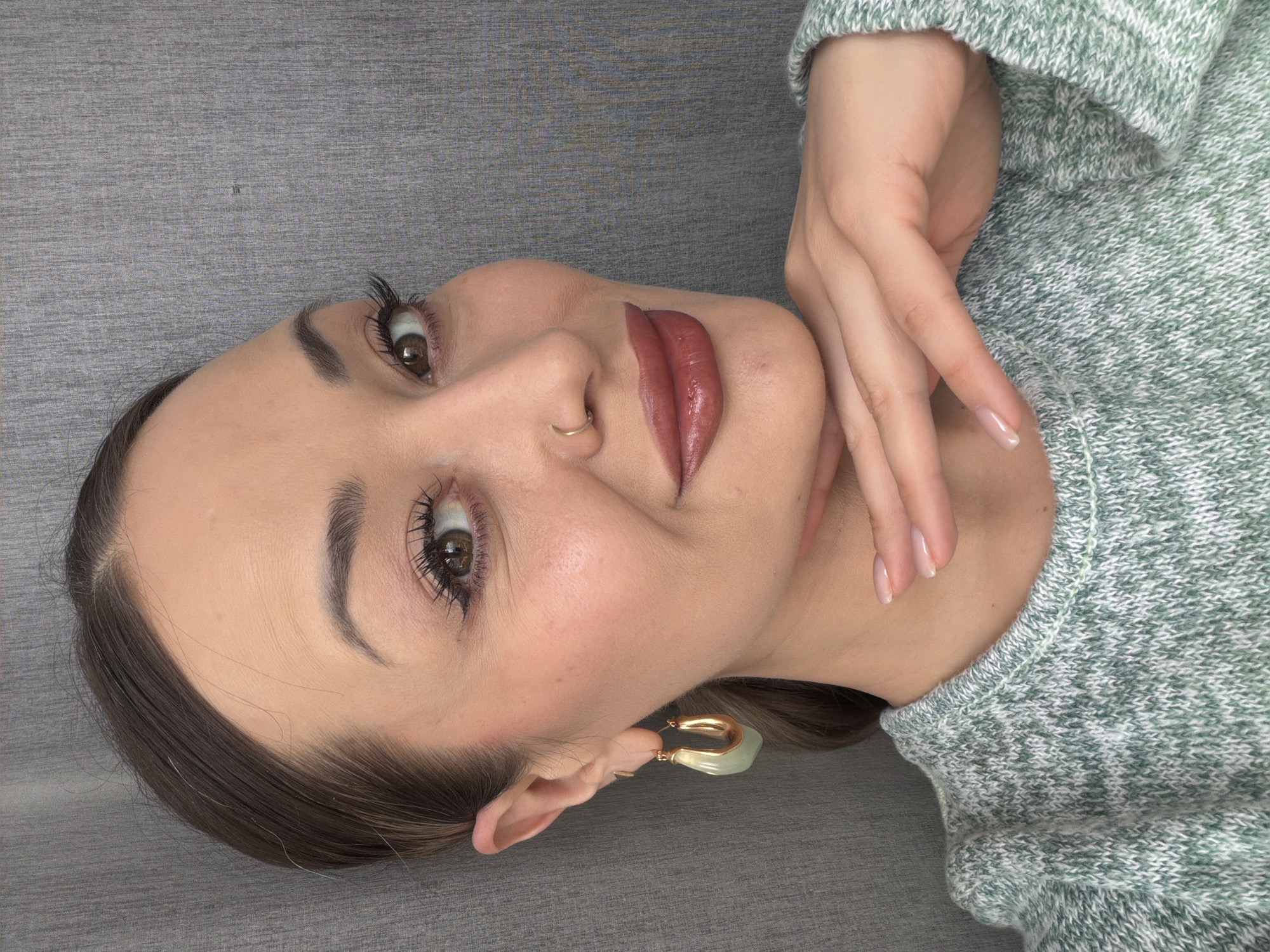 A woman with fair skin, brown hair, and brown eyes is lying down on a gray surface, possibly a couch or bed. She is wearing a light green sweater with a white speckled pattern. Her right hand is raised near her face, fingers gently curved. She has a gold hoop earring on her left ear and a gold ring in her left nostril. Her lips are painted with a glossy, brownish-red lipstick. The location appears to be indoors, but there is no information indicating that this image was taken in Krasnoyarsk, Russia.
