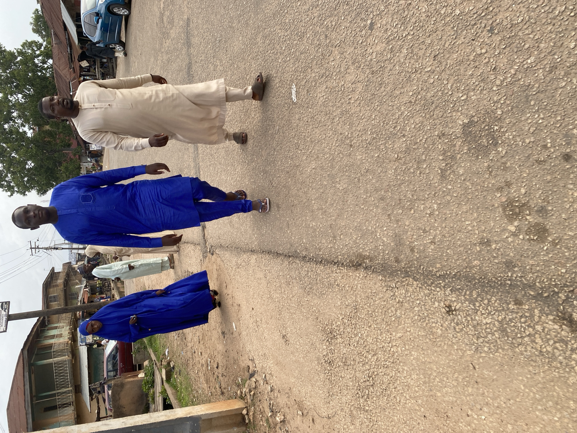 A street scene in Jos, Nigeria, features a woman in a blue hijab and a long blue dress walking to the left, facing the camera. She is followed by two men in traditional clothing. The man closest to her is wearing a light beige tunic and pants, walking towards the right. The man behind him is wearing a bright blue tunic and pants, also walking towards the right. The background shows a street with buildings, including a two-story structure with a balcony, and a blue car parked on the right side of the road. Trees are visible in the distance. The ground is a paved road.