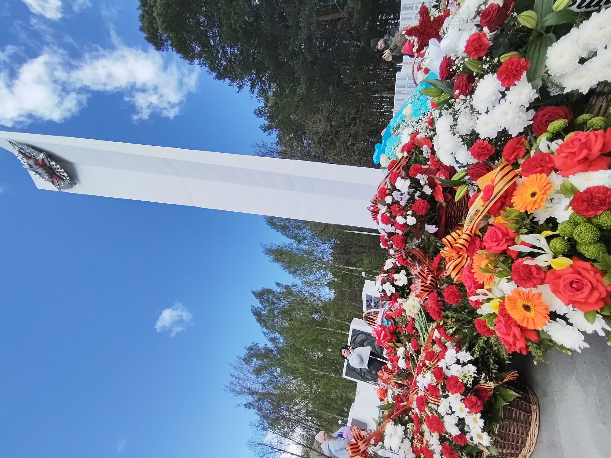 The image shows a monument in the city of Chaikovsky, Russia. The monument is a tall, white structure with a metal emblem at the top. There are several flower arrangements at the base of the monument, including red, white, and orange flowers. The flowers are arranged in baskets and tied with orange and black ribbons. The sky is blue with a few clouds. There are trees in the background.