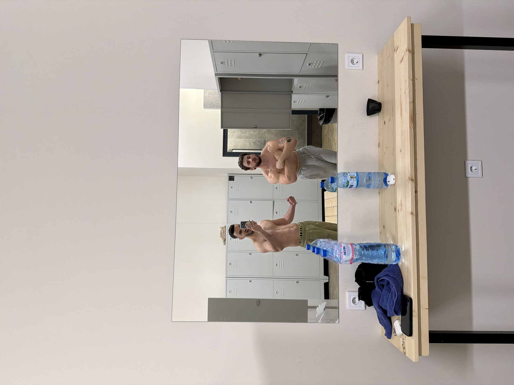 The image captures a scene in what appears to be a gym locker room, oriented with a large mirror on the left and a wooden shelf on the right. In the mirror's reflection, two shirtless men are visible, both posing and flexing their muscles. The man closer to the viewer in the reflection wears grey sweatpants and has a tattoo on his left bicep, looking intently at himself. Further back in the reflection, a second man, wearing olive green pants, is holding a smartphone to take a selfie while also flexing. The reflection also shows rows of grey metal lockers in the background, characteristic of a changing facility.
To the right, a natural wood shelf is mounted against a light-colored wall, supported by a black metal beam. On the shelf, there are two clear plastic water bottles, both clearly displaying "Devin" branding. Alongside these are a folded blue towel, a black t-shirt, a black smartphone, and a set of keys attached to a white wireless earbud case. Two white electrical outlets are visible on the wall; one is positioned above the shelf near the mirror, and another is further to the right. A small black hook or fixture is also mounted on the wall above the shelf. The scene suggests a post-workout moment, with the individuals admiring their physique in the mirror. The "Devin" water bottles specifically tie the location to Bulgaria, consistent with the Plovdiv context.