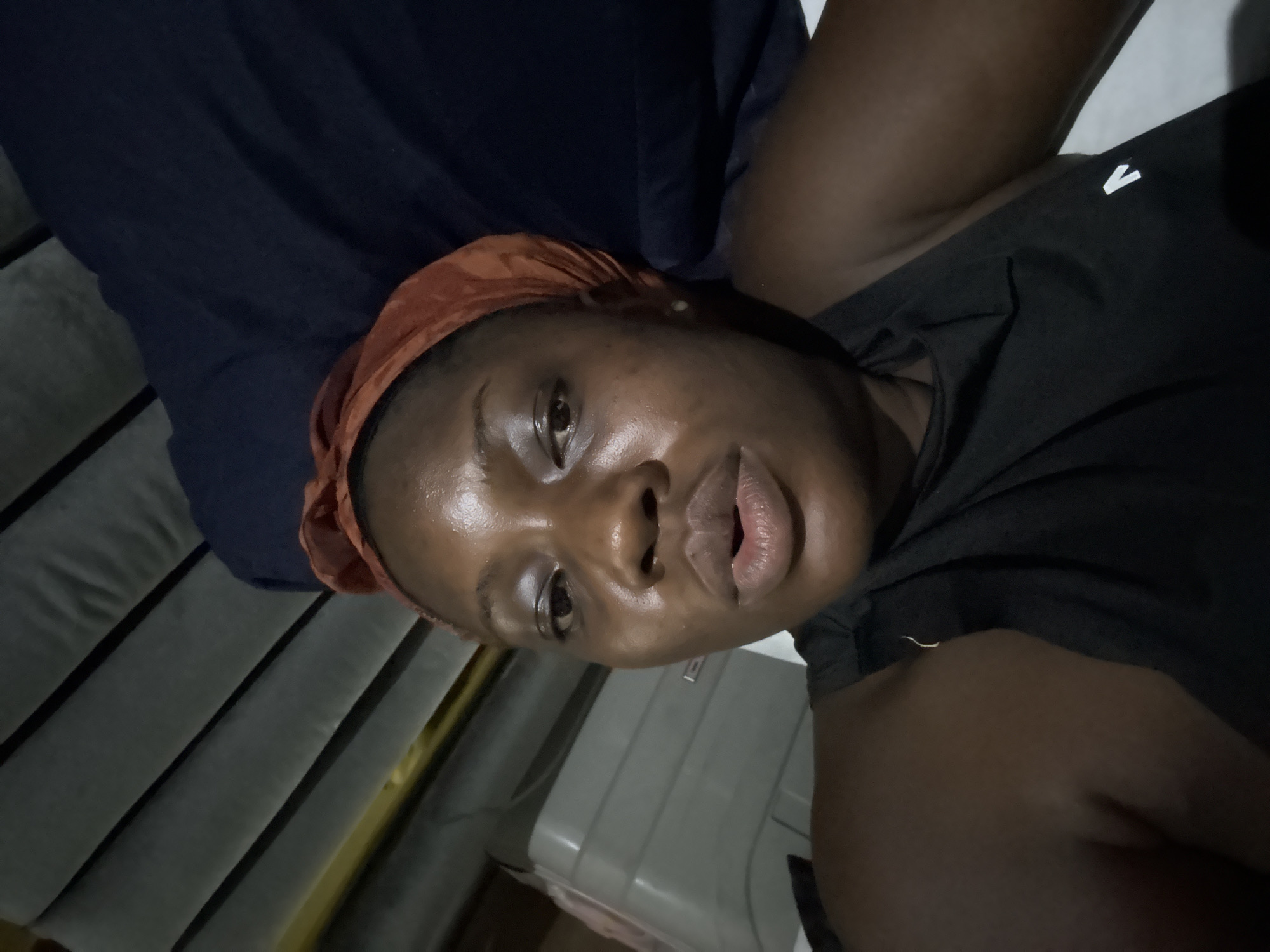 The image shows a person with brown skin and an orange headscarf. The person is wearing a black t-shirt with a white logo. The background consists of a blue cushion and a gray ribbed surface. The location in the city of Iddo Tudunwada, Nigeria is not discernible from the image.