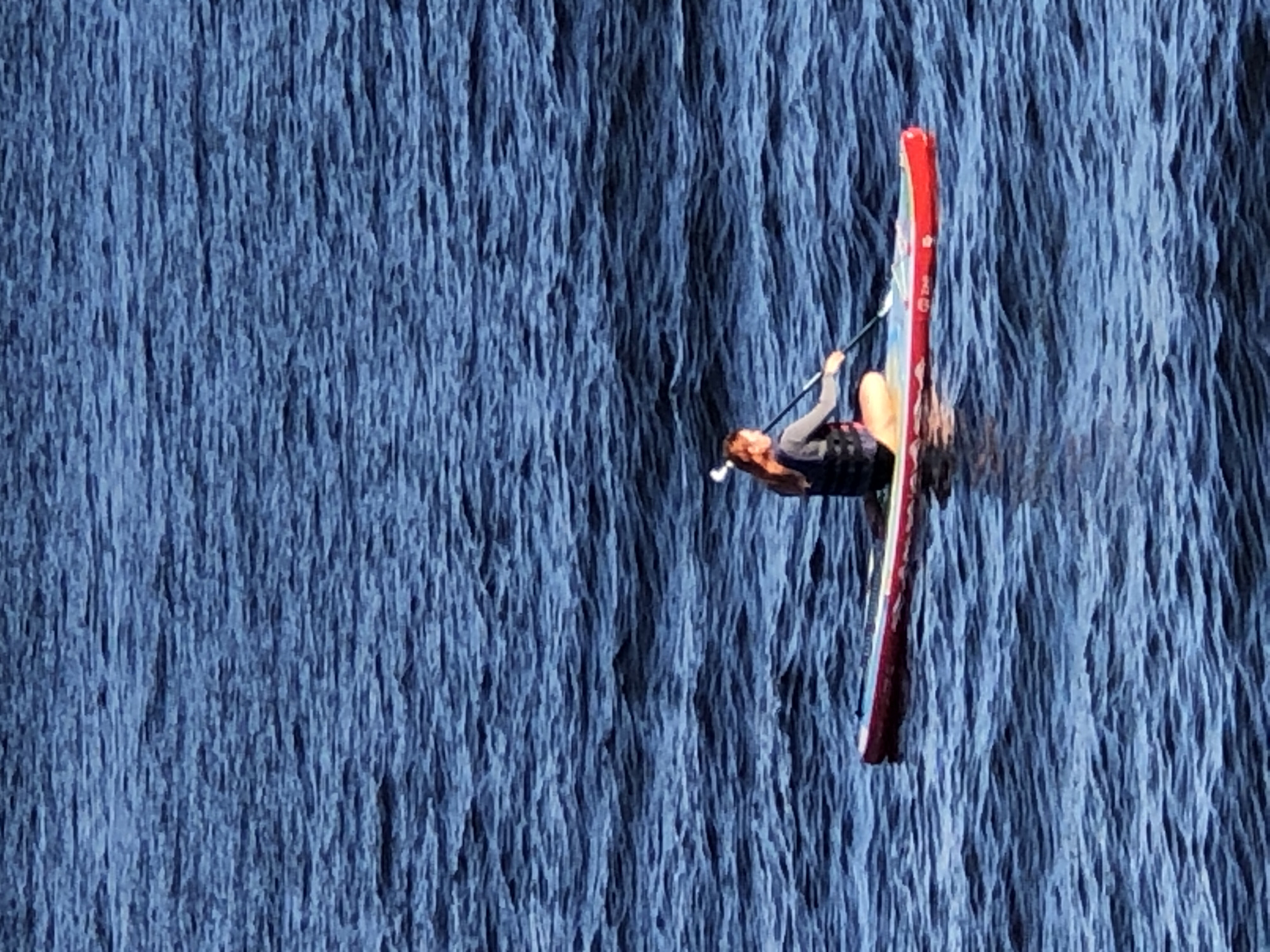 A person with auburn hair is paddleboarding on a body of water. They are wearing a life vest and are seated on a paddleboard with red, white, and blue accents. The person is holding a paddle and is positioned in the center of the frame. The water is dark blue and choppy, with small waves visible throughout the image. The scene appears to be outdoors.