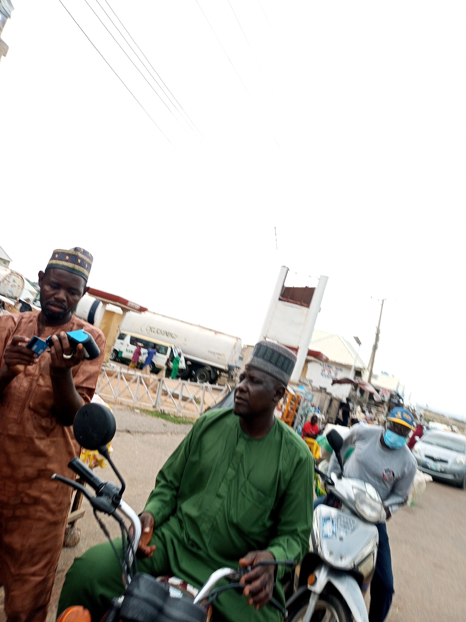 The image depicts a street scene in Gombe, Nigeria, with a focus on a group of men and their motorbikes. In the foreground, a man wearing a brown traditional outfit and a patterned cap is holding a blue card and a black object, possibly a mobile phone. To his right, another man in a green traditional outfit is seated on a motorbike, looking to the side. Behind him, a third man, wearing a grey long-sleeved shirt, dark pants, and a blue face mask, is also on a motorbike. He has a yellow and blue cap on his head. In the background, there are vehicles, including a large white tanker truck, a white van, and a silver car, suggesting a commercial or industrial area. The environment appears to be a street with various structures and stalls, indicating a busy urban setting. The overall lighting suggests it is daytime, with overcast skies.