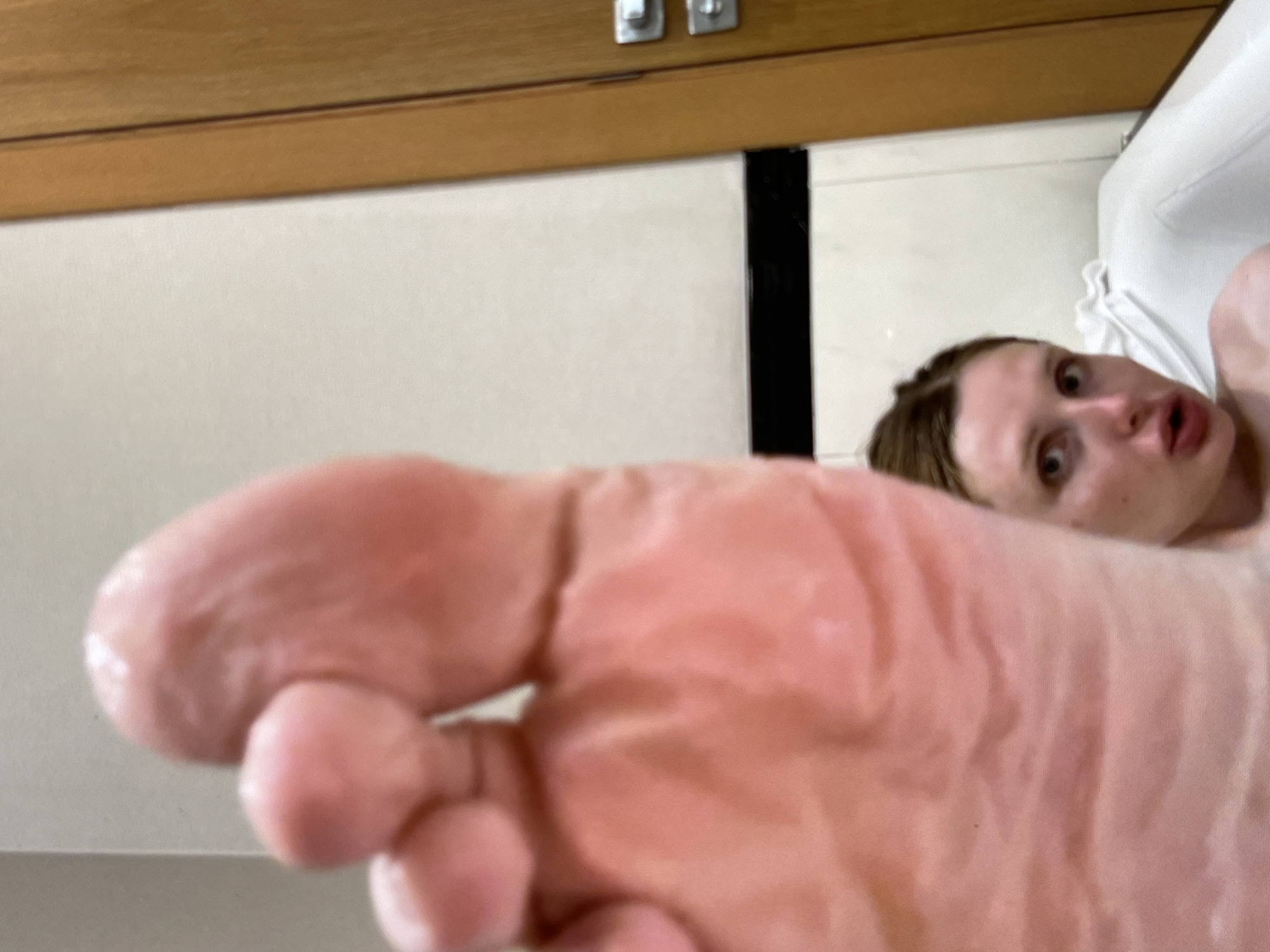 This media file shows a close-up of a person's foot, with the person's face blurred in the background. The foot appears to be pinkish-orange and has visible wrinkles. The person's face is partially visible, with wide eyes and an open mouth. They have light-colored hair. The background includes what appears to be a light-colored wall or tiled surface, and a wooden element at the top, possibly a cabinet or shelf. There is also a black vertical stripe in the background. The lighting suggests an indoor setting. The overall impression is of a selfie or a photograph taken from a low angle, focusing on the foot with the person's face serving as a secondary, out-of-focus element. The emotion conveyed by the visible part of the face is one of surprise or excitement. There is no visible text in the image.