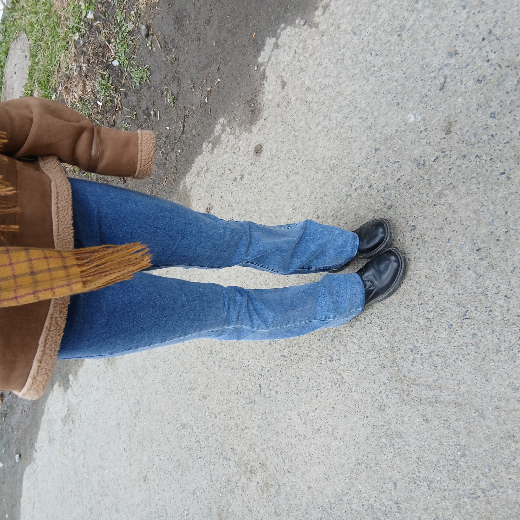 The image provides a top-down view of a person standing or walking outdoors. The individual is wearing a brown, faux fur-lined jacket or coat, blue jeans, and black, slip-on shoes. A plaid scarf, primarily yellow with brown and possibly reddish patterns, is visible draped around the upper body, with fringed ends extending towards the bottom of the frame.
The ground surface is a mix of textures. On the right side of the frame, and under the majority of the person's stance, is light grey concrete pavement, typical of a sidewalk or path, with visible aggregate. On the left side of the frame, there's an area of darker, disturbed earth with sparse patches of green and dry grass, along with some small twigs and debris. This dirt patch appears somewhat damp or muddy, contrasting with the drier concrete.
The scene depicts a casual outdoor moment. The natural, even lighting suggests it is daytime, possibly overcast, as there are no strong, distinct shadows. The warm attire, including a jacket and scarf, implies cooler weather conditions. There is no visible text in the image. The environment suggests an urban or suburban setting, possibly a pathway adjacent to a natural strip of ground.