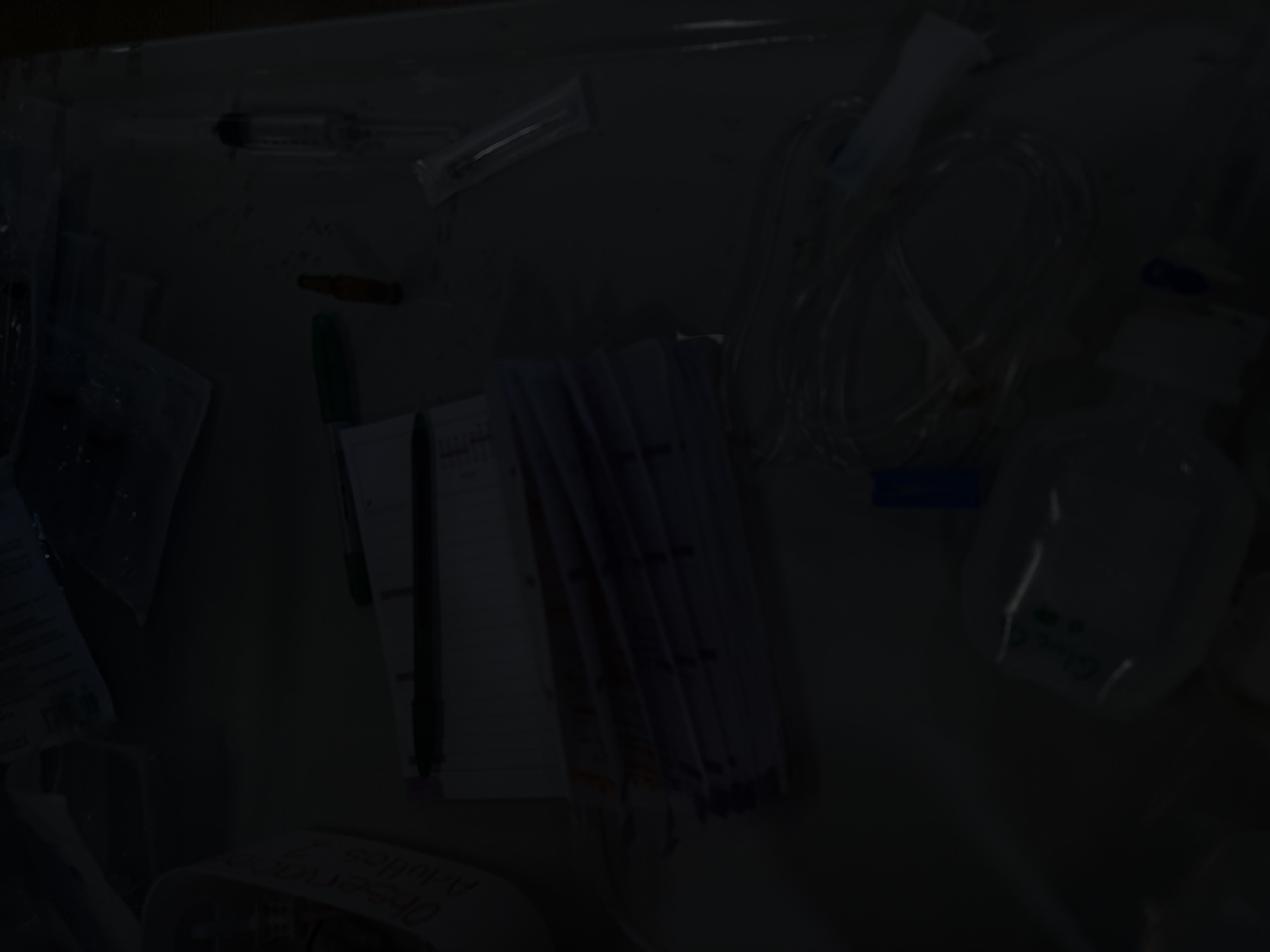 The image shows a dimly lit medical setting, possibly a hospital room. There are various medical supplies scattered across a surface, including syringes, needles, intravenous tubing, and an IV fluid bag. Medical paperwork and writing instruments are also present. The overall scene suggests a medical examination or treatment area, although the poor lighting makes detailed identification difficult. There is no information to suggest that this photo was taken in Puebla, Mexico.