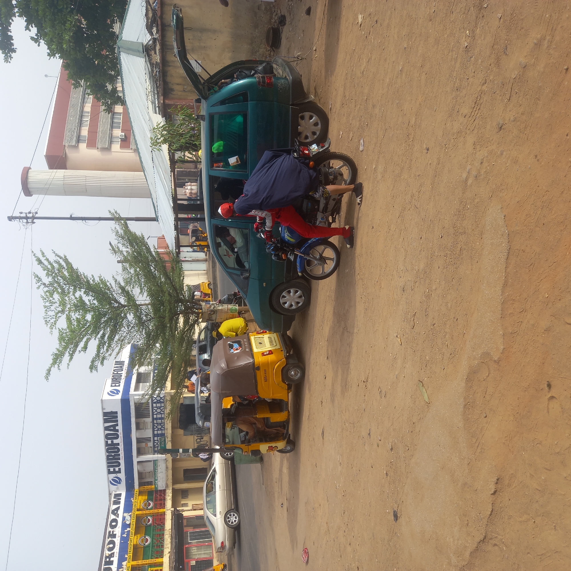 The image depicts a street scene in what appears to be the city of Sardauna, Nigeria, characterized by a mix of vehicles and infrastructure. A green hatchback car is parked on a dirt area, with its trunk open. Next to it, a motorcyclist, clad in red trousers and a red cap, is interacting with the open trunk. A yellow tricycle, presumably a local transportation vehicle, is parked further down the road. In the background, there is a building with a sign that reads "EUROFOAM." Other vehicles are visible, as well as pedestrian activity. A cylindrical structure is visible in the background. The overall environment suggests a semi-urban or urban setting with a dusty, unpaved roadside.