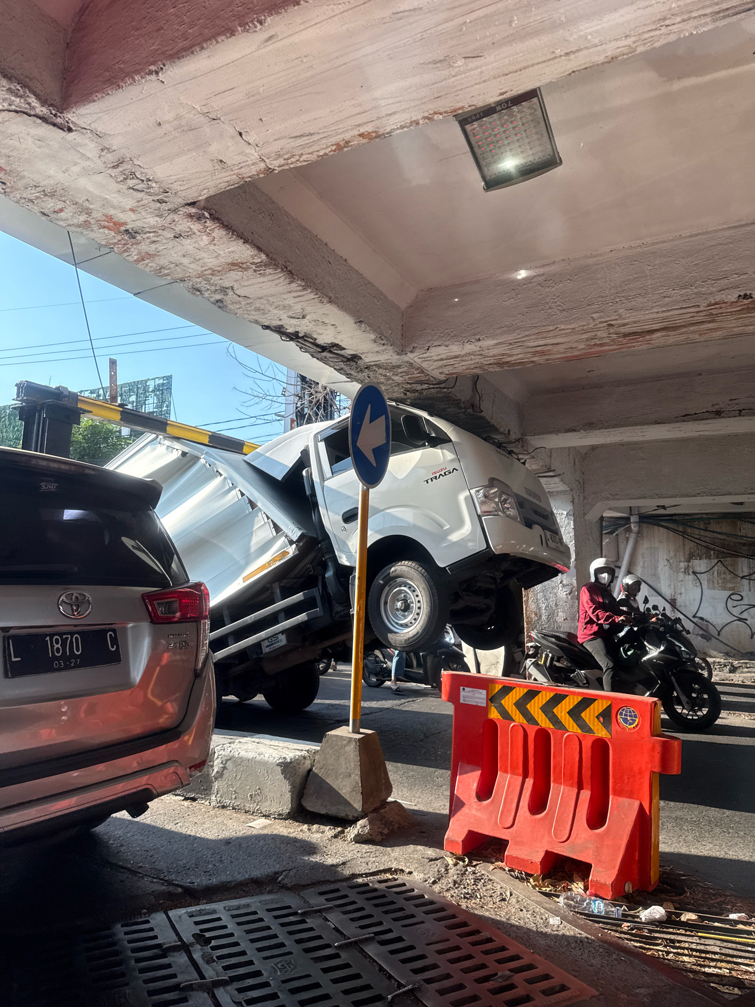 A white truck is wedged under a concrete overpass in Surabaya, Indonesia, its front end lifted high and tilted. The truck's bed is also significantly damaged and bent. A silver SUV is parked behind the truck, facing the same direction. Two people on a black motorcycle, wearing helmets, are stopped behind the SUV. A bright orange traffic barrier is positioned on the right side of the road, near the motorcycle. The overpass shows signs of damage, with visible cracks and exposed rebar in the concrete. The sky is clear and blue.