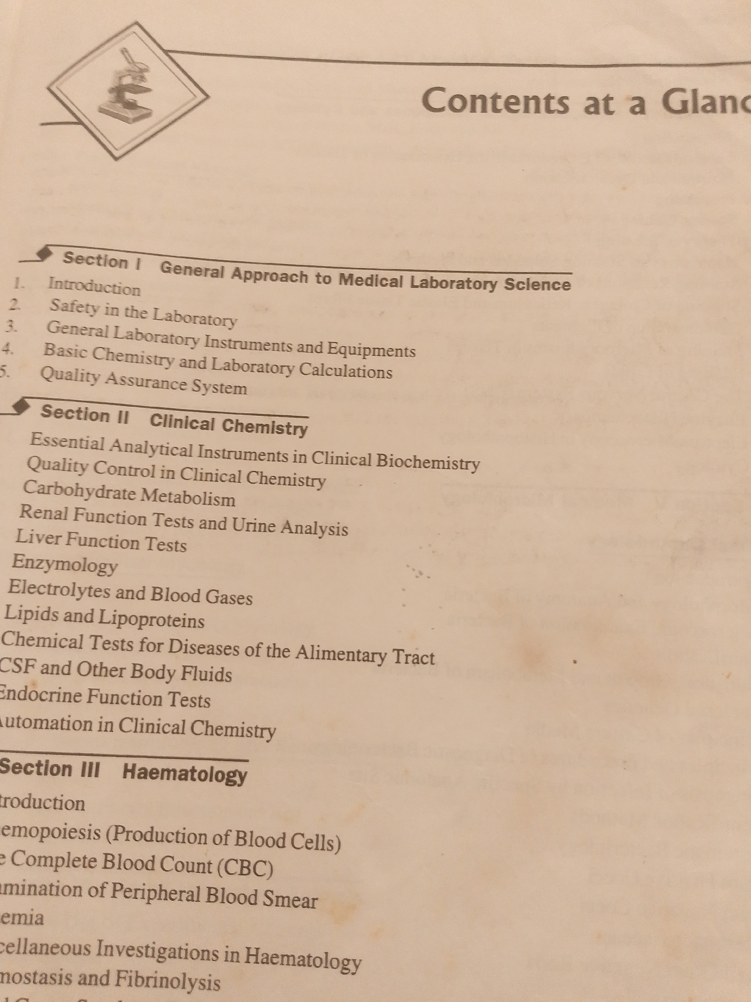 The image displays a table of contents from a book, likely a medical or scientific textbook, as indicated by the section titles such as "General Approach to Medical Laboratory Science," "Clinical Chemistry," and "Haematology." The page is slightly yellowed and shows signs of age.
The visible text includes the heading "Contents at a Glance," followed by three main sections. Section I details topics like "Introduction," "Safety in the Laboratory," and "Quality Assurance System." Section II, "Clinical Chemistry," lists subtopics such as "Essential Analytical Instruments in Clinical Biochemistry," "Carbohydrate Metabolism," and "Liver Function Tests." Section III, "Haematology," includes entries like "Haemopoiesis (Production of Blood Cells)" and "Complete Blood Count (CBC)."
In the top left corner, there is a stylized icon of a microscope within a diamond shape, further reinforcing the scientific nature of the content. The overall setting appears to be a book page. There are no people, animals, or depictions of natural scenery. The image is well-lit, with no indication of time of day or weather. The location context of Jalingo, Nigeria, does not directly inform the visual content of the image itself, other than suggesting the origin or target audience of the document. The tone is purely informational and academic.