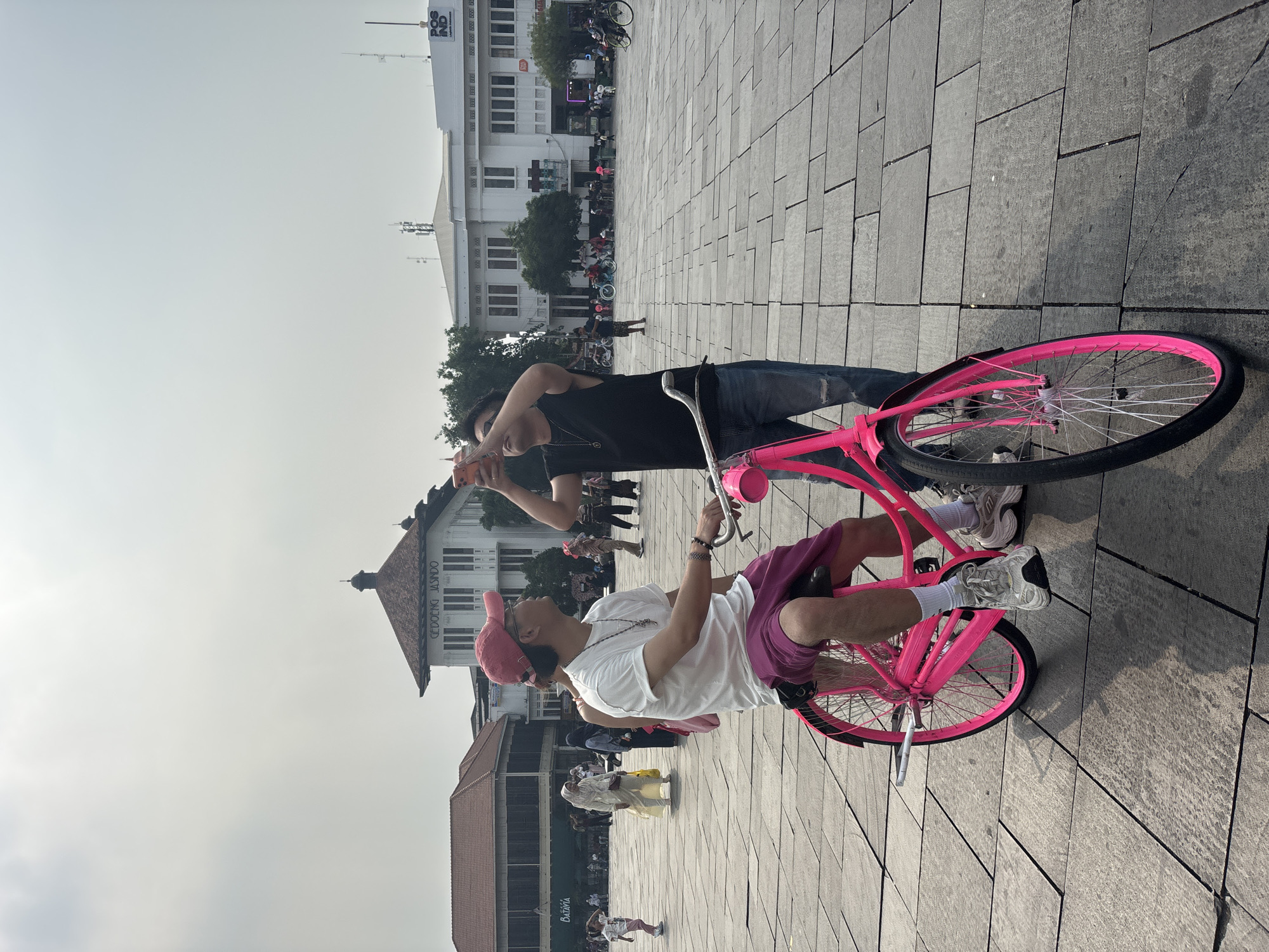 In a paved square in Kota Tua, West Jakarta, Indonesia, two young men are in the foreground. The man on the left is seated on a vibrant pink bicycle, wearing a white t-shirt, purple shorts, a red baseball cap, and silver athletic shoes. The man on the right, standing and holding a mobile phone, is taking a photo or video of the man on the bicycle. He is wearing a black sleeveless shirt, ripped blue jeans, and dark shoes. The bicycle has a silver frame and pink wheels. In the background, several other people are visible, some walking and others on bicycles, including a person in a yellow hijab and a person in a white hijab. The architecture in the background suggests a historic district, with colonial-era buildings. The sky is overcast.