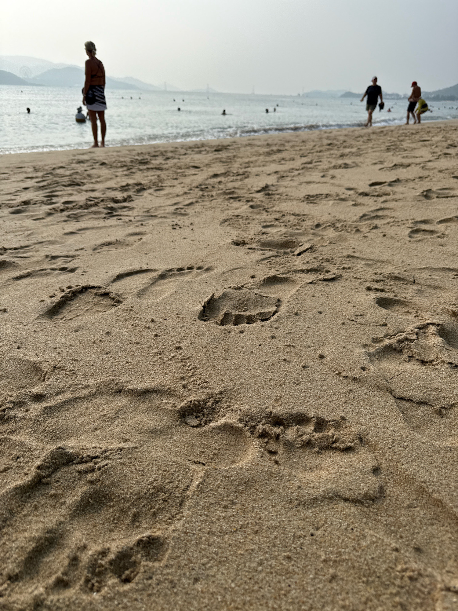 The image captures a low-angle view of a sandy beach, with footprints prominently visible in the foreground. The sand is a light brown color, with some areas showing darker, damp patches. In the background, the ocean meets the shore with gentle waves. Several figures are present on the beach and in the water, appearing as silhouettes against the hazy sky. One person stands closer to the water's edge, facing away from the camera, wearing a dark top and a striped skirt. Further out in the water, other individuals can be seen wading or swimming. The distant shoreline is lined with hills and structures, suggesting an urban or semi-urban coastal environment. The overall atmosphere is calm and serene, characteristic of a beach scene. This scene is likely located on the coast of Nha Trang, Vietnam, a city known for its extensive beaches and scenic bay.