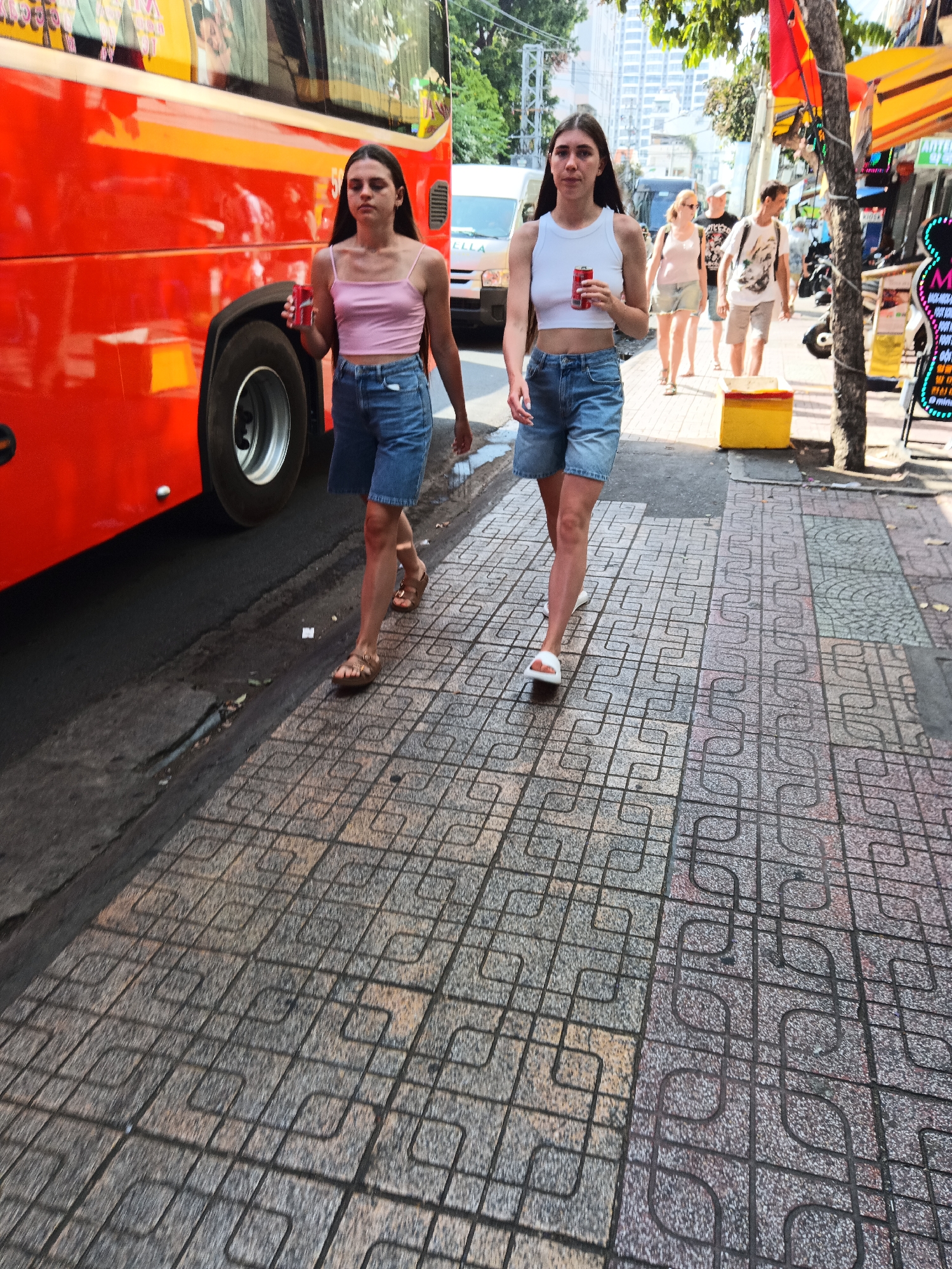 Two young women, one in a pink spaghetti-strap crop top and denim shorts, the other in a white tank top and denim shorts, walk on a tiled sidewalk adjacent to a large red bus and a busy street. The woman in pink holds a red can in her right hand, while the woman in white holds a similar can in her left. Both are wearing sandals. In the background, other pedestrians and vehicles are visible, including a white van. The scene appears to be in Nha Trang, Vietnam, indicated by the architecture and signage, possibly along a street with shops. The lighting suggests it is daytime.