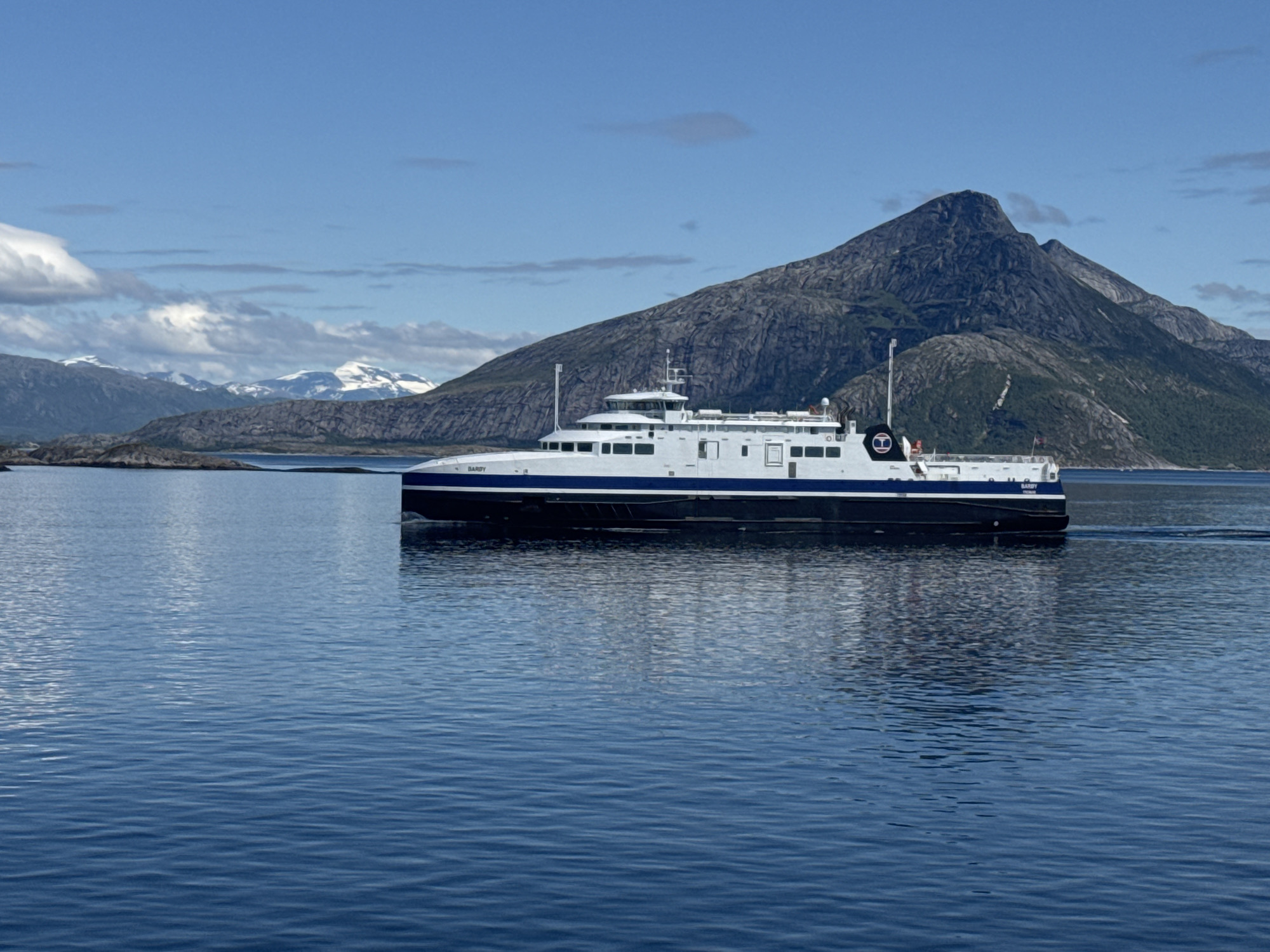A large white and dark blue ferry boat is sailing across calm blue water. The ferry has several decks and windows, with a tall white structure at the front. There is text visible on the side of the ferry that reads "BARDY". In the background, there are rugged, rocky mountains with some snow-capped peaks. The sky is a clear blue with some scattered white clouds. The scene appears to be set during the daytime, under clear weather conditions. The overall impression is one of serene travel in a scenic, mountainous coastal environment.