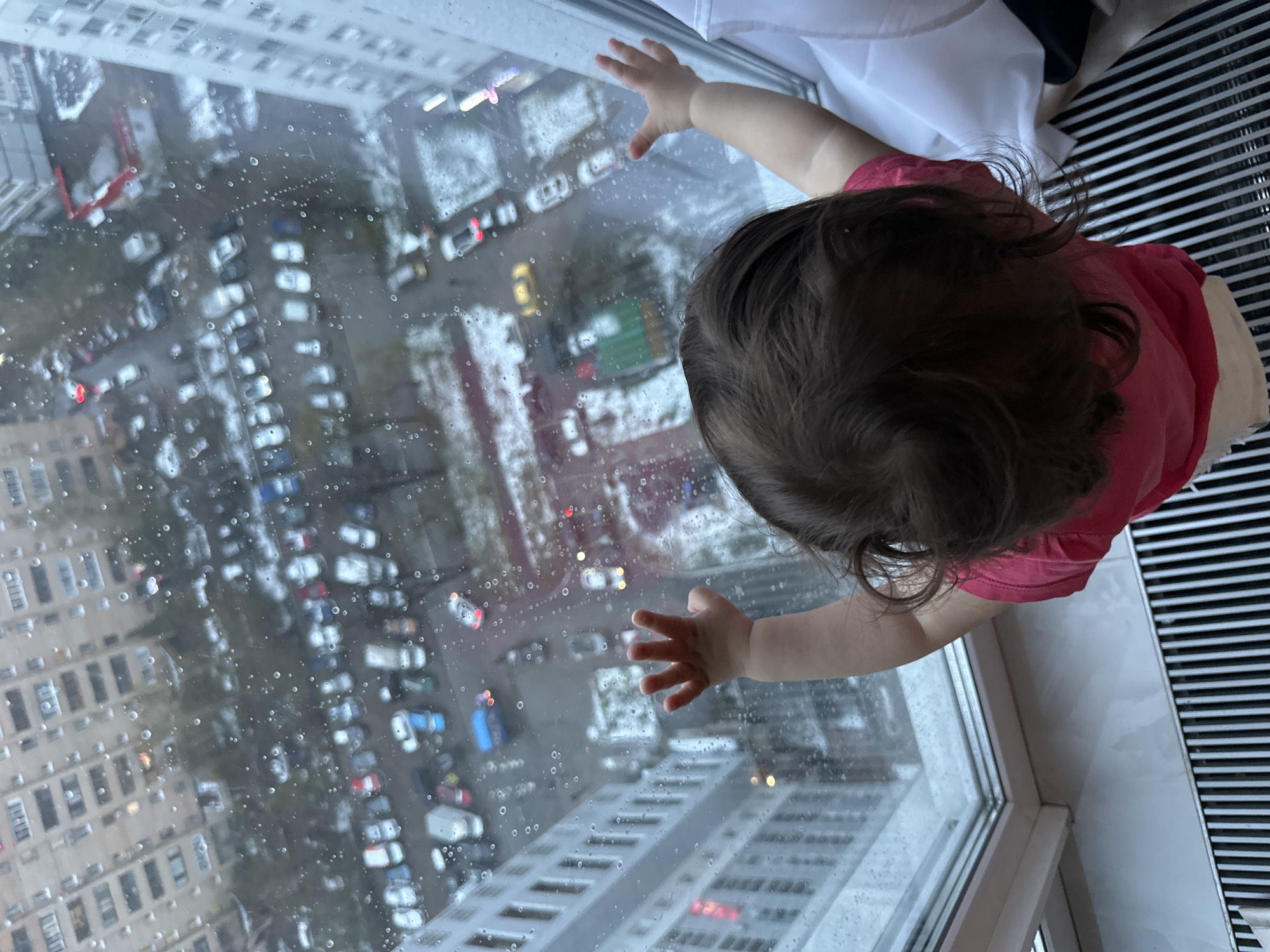 The image presents a high-angle, slightly rotated view of a young child looking out a window onto an urban scene. The child, seen from the back, has dark, curly hair and wears a pink or reddish t-shirt. Their small hands are extended and gently pressed against the rain-streaked window pane, conveying a sense of curiosity or engagement with the outside.
Indoors, the child is positioned on what appears to be a white window sill, with a black and white striped grate, likely a heating vent or radiator cover, visible to the right.
The view outside is a dense urban landscape, with numerous multi-story residential buildings occupying the background. Streets below are lined with parked cars, and other vehicles can be seen moving. The ground exhibits patches of white, indicating snow or slush, and the window is covered in prominent raindrops, confirming that it is a cold, wet, and overcast day. Some car lights are on, and lights are visible in some building windows, suggesting it might be late afternoon or early evening. The scene depicts a typical busy city environment, consistent with the provided location context of Moscow, Russia. No visible text is present in the image.