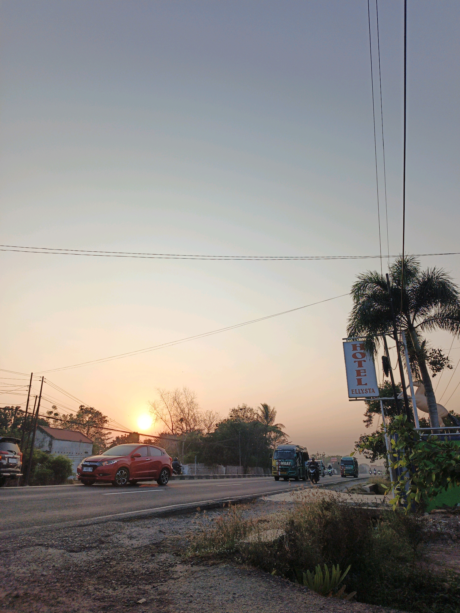 The image depicts a roadside scene in Krimun, Indonesia, during sunset. The sky is a soft gradient of pale blue and orange, with the sun visible as a bright orb low on the horizon, casting a warm glow. A red SUV is driving on the asphalt road, with other vehicles, including buses and motorcycles, also present. On the right side of the road, a sign for "HOTEL ELLXYSTA" is displayed, attached to a pole and partially obscured by a palm tree. The foreground shows a gravelly embankment with dry grass and vegetation. Power lines crisscross the sky, supported by utility poles. The overall atmosphere is one of a tranquil evening on a busy road.