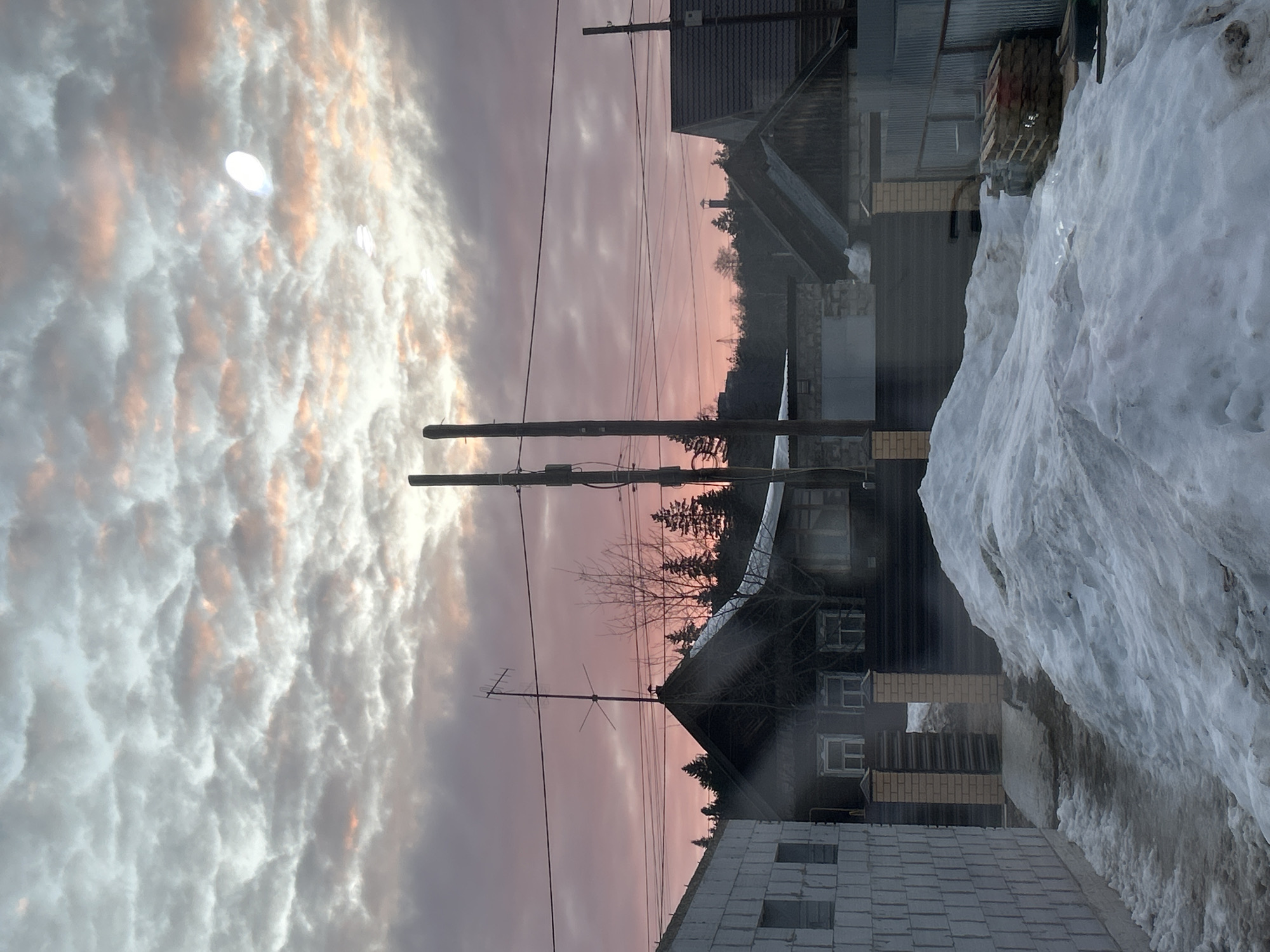 This image depicts a residential scene in Chaikovsky, Russia, during what appears to be either sunrise or sunset, given the atmospheric lighting. The sky is largely overcast with stratified clouds displaying hues of grey, white, pink, and orange, particularly closer to the horizon. A bright, diffused white oval light source, possibly the sun, moon, or a lens flare, is visible in the upper right portion of the sky.
In the foreground, a significant amount of snow covers the ground, forming undulating mounds. A dark, partially cleared path, possibly wet or muddy, leads from the lower center into the scene. To the left of this path is a partially constructed building made of light-colored cinder blocks, with exposed walls and rectangular openings for future windows. To the right of the path is a large snowdrift. Further right, near a dark fence with brick pillars, a stack of wooden pallets and other construction materials are visible.
The midground features two tall, dark utility poles with multiple power lines running horizontally across the frame. Beyond these poles, several residential buildings with dark roofs are visible. One prominent dark-colored wooden house with white-framed windows and a television antenna mounted on its pitched roof stands out towards the left. Bare deciduous trees are interspersed among the buildings.
The background consists of a dense line of dark coniferous trees forming a horizon. Above these trees, a faint, dark, cylindrical structure, possibly a chimney or a distant tower, is visible on the far horizon. No individuals or active human interactions are present in the scene. The overall environment suggests a cold, winter setting with ongoing construction in a semi-rural or suburban residential area.
