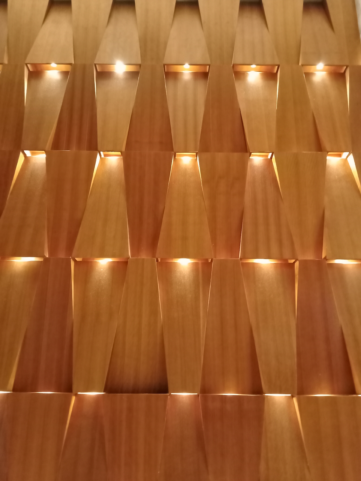 This media file captures a close-up of a modern, intricately designed wooden wall paneling system, likely located indoors in Nha Trang, Vietnam. The wall is composed of numerous individual wooden panels arranged in a repeating geometric pattern, creating a textured, three-dimensional surface.
The panels are made of a light to medium-toned wood, showcasing natural grain variations. They are angled and offset, forming horizontal recesses at regular intervals across the wall. Within these recesses, small, bright light sources (likely LEDs) are embedded, casting a warm, inviting glow both upwards and downwards onto the wooden surfaces. This integrated lighting highlights the architectural contours of the panels, creating a dynamic interplay of light and shadow.
There are no people or discernible activities taking place. The scene focuses solely on the decorative wall. The warm, ambient lighting suggests an interior setting, possibly in the evening or a space designed for a cozy atmosphere, such as a hotel lobby, restaurant, or contemporary living area. The design conveys a sense of elegance, modernity, and warmth. No text is visible in the image.