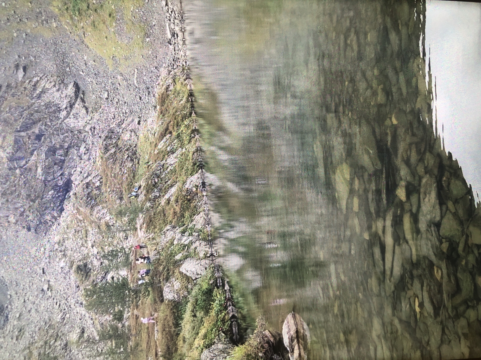 A landscape photograph shows a rugged, rocky mountainside partially obscured by water. On the left, a steep slope covered in loose gravel and large boulders ascends. Vegetation, including grasses and small trees, clings to the rock face. A group of people are visible on a narrow trail or ledge along the slope. The right side of the image is dominated by water, possibly a lake or river, reflecting the mountainside. The water is murky and indistinct, creating a distorted and partially obscured view of the submerged rocks. The location and city cannot be determined based on the image.