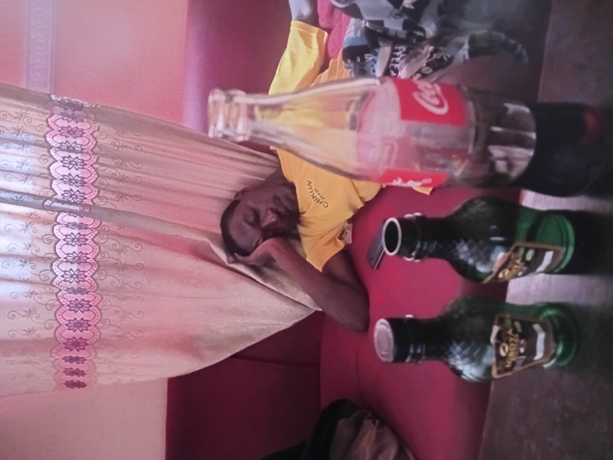 A man is asleep on a red sofa. He is wearing a yellow t-shirt with the word "CHIKUN" written on it. In front of him are three bottles on a table. Two of the bottles are dark green, and one is a clear Coca-Cola bottle. The room has a pink curtain with a floral pattern. The scene appears to be in an indoor setting, likely a living room, in Jos, Nigeria.