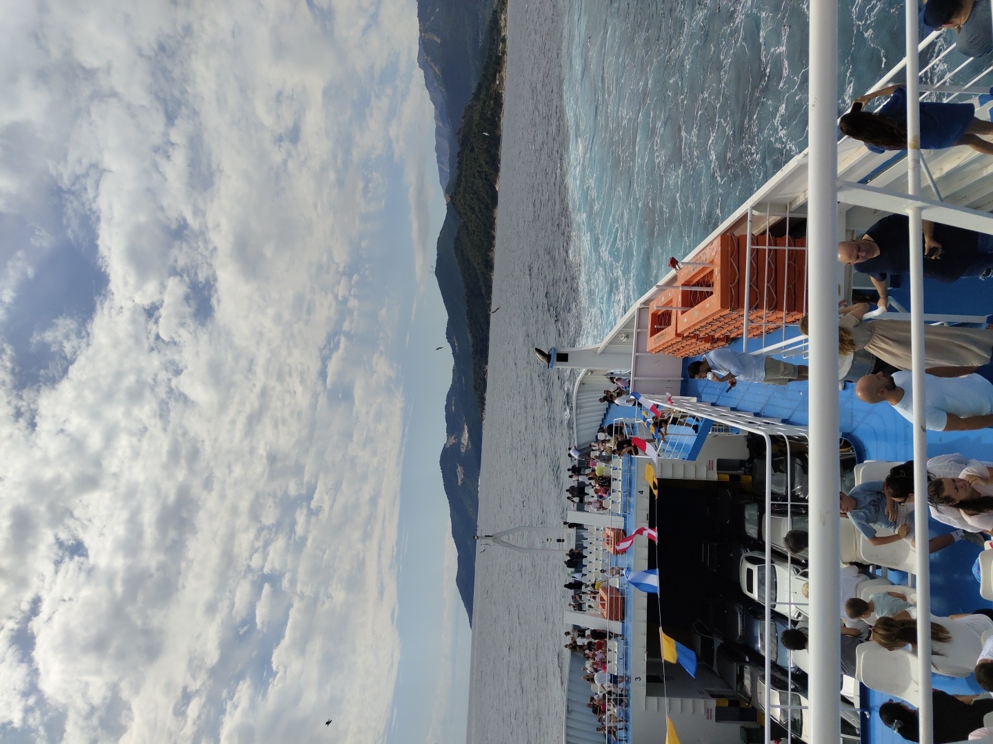 This image captures a sunny day scene on a passenger ferry at sea, likely departing from or arriving at the port of Thassos, Greece. The foreground features a white railing of the ferry, behind which passengers are seated on white chairs on the blue deck. Further back, the ferry's open car deck is visible, filled with parked cars. On the upper deck, many passengers are standing and sitting, some looking out at the sea. Orange life rafts are stacked on the side of the ferry. In the background, a coastline with green, hilly terrain and mountains meets the deep blue sea. The sky is partly cloudy with scattered white clouds against a bright blue sky. Several birds are visible in flight. The ferry appears to be in motion, with white wakes churning in the water.