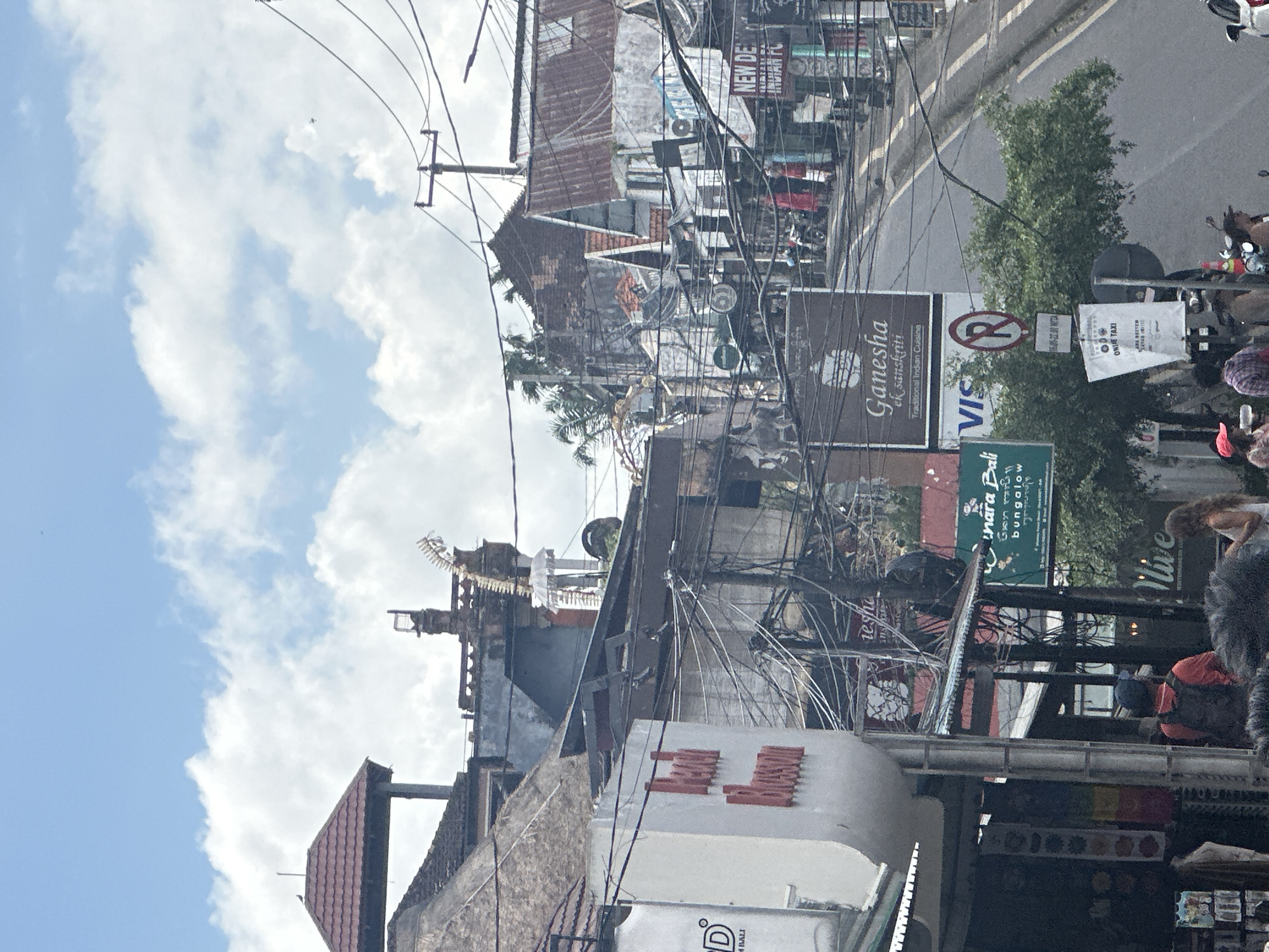 The image depicts a street scene in Ubud District, Indonesia, characterized by a dense network of overhead electrical wires crisscrossing above buildings. The architecture features traditional Balinese elements, including pitched roofs and ornate details, with some structures appearing aged or weathered. Various signs are visible, advertising businesses such as "Hotel Blossom," "Ganesha Traditional Indian Cuisine," and "Sunara Bali Bungalow." A "No Parking" sign is also present. Several people are seen on the street, some walking and others on motorbikes. The overall impression is a busy urban environment with a mix of commercial activity and local life.