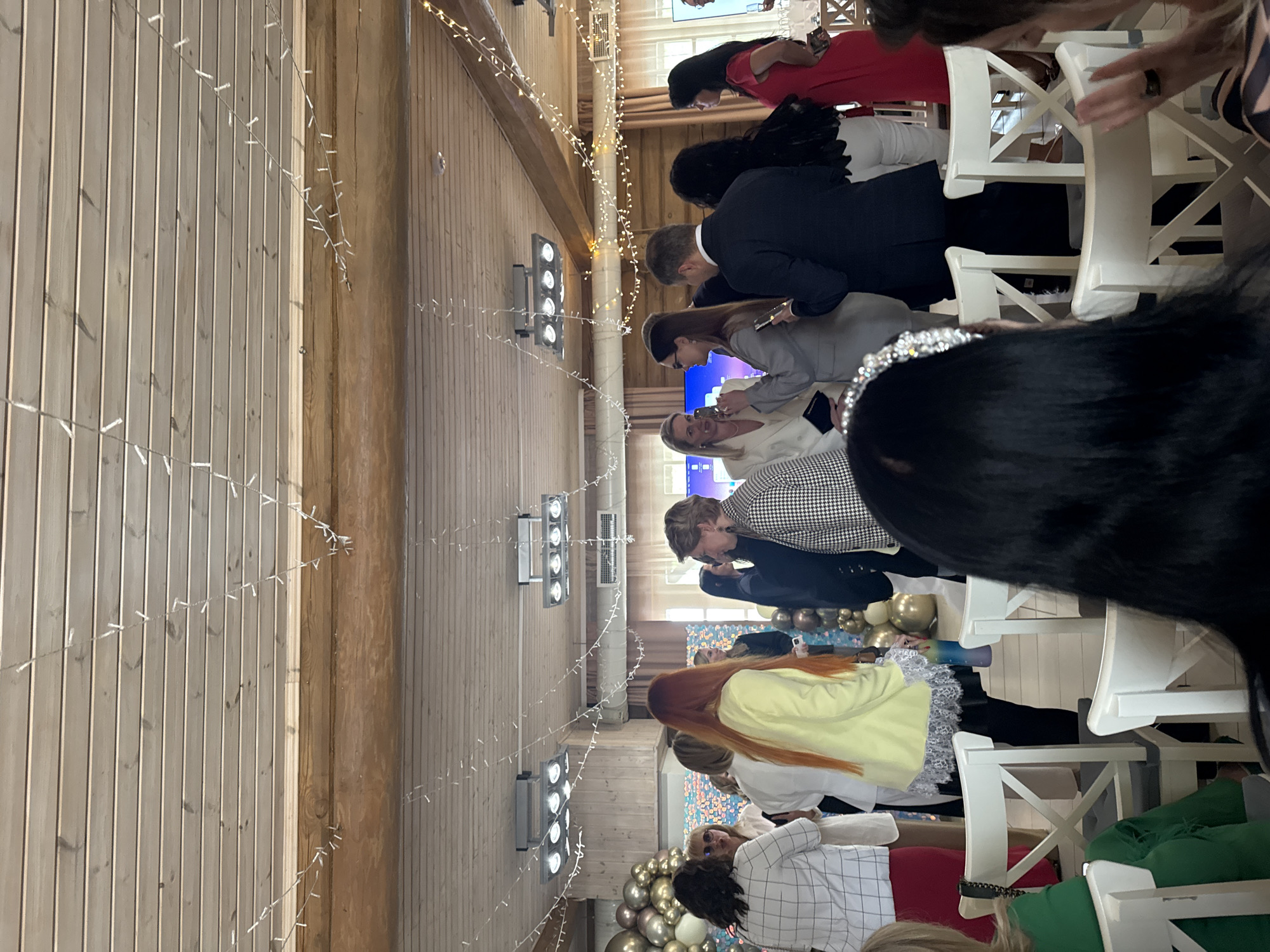 The image shows the interior of a room, likely a venue for an event, in Lilaste, Latvia. The walls are paneled with light-colored wood, adorned with strings of lights. People are seated in white chairs, facing a central area. They are dressed in a variety of formal and semi-formal attire. Some individuals appear to be looking at a screen or stage. Decorations include balloons.