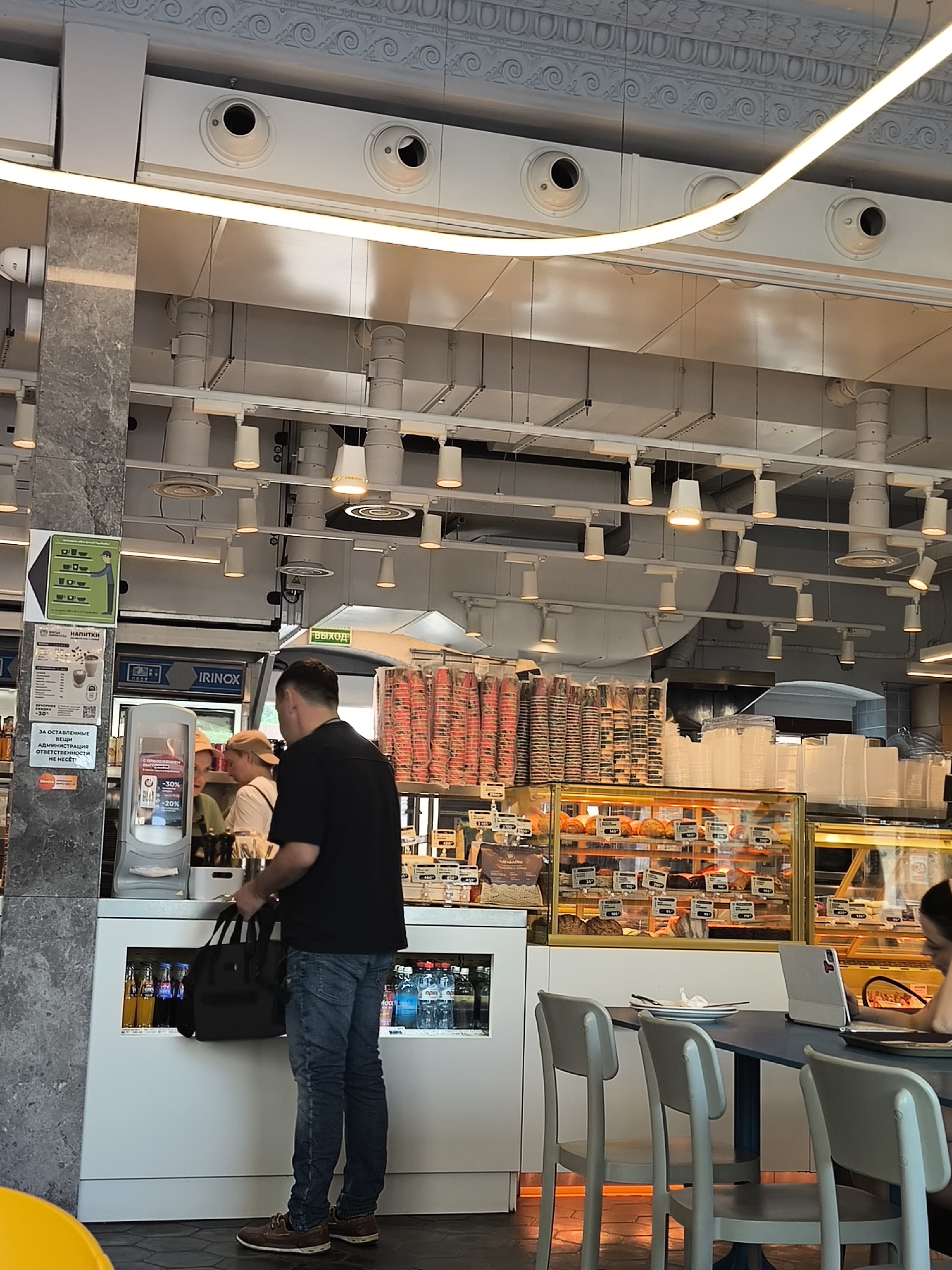 The image depicts the interior of a café or bakery, likely located in Unknown, Unknown. The scene is bustling with activity. A man, dressed in a black shirt and blue jeans, is seen at the counter, seemingly in the process of making a purchase, as indicated by the bag he is holding. In the background, a woman is seated at a table, occupied with a laptop. Display cases filled with pastries are visible, suggesting the café's offerings. The café is equipped with various lighting fixtures and ventilation systems. Signage with text, presumably in Russian, is displayed on a pillar. The general atmosphere of the café is modern and well-lit.