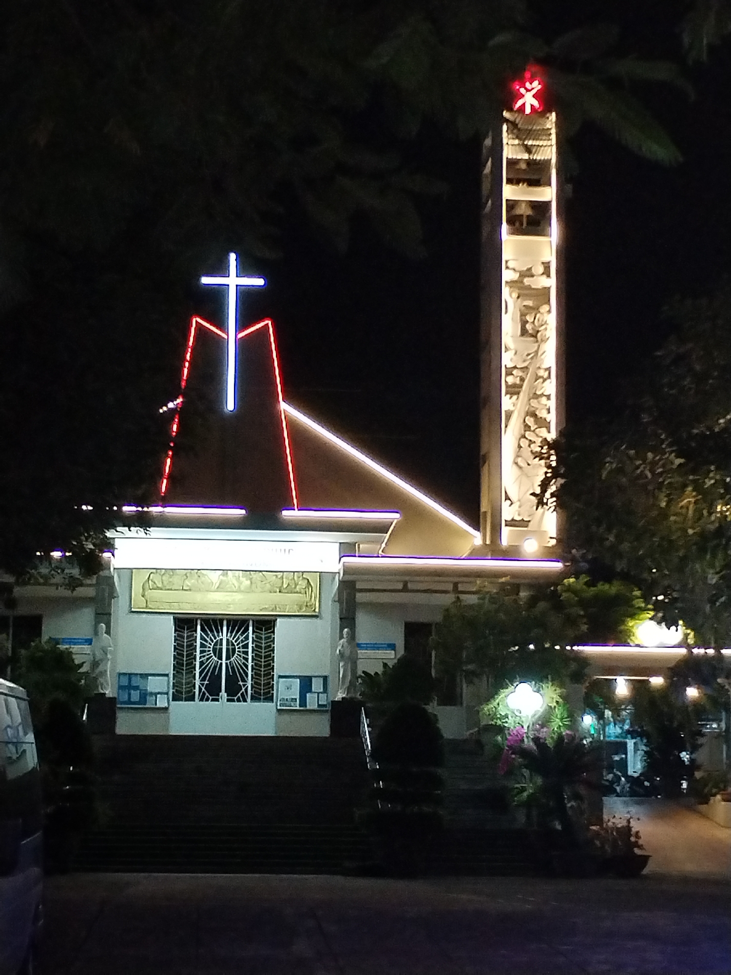 At night, a church in Bac Nha Trang Ward, Vietnam, is illuminated. A tall bell tower stands to the right of the building, topped with a red symbol. A blue cross is prominently displayed on the front of the church, outlined in red neon. White lights run along the roofline of the structure. Steps lead up to the building's entrance. The Last Supper is depicted above the entrance. Two statues flank the doors, and bulletin boards are visible on either side. Lush trees surround the building. A car is parked to the left of the image. A streetlight illuminates landscaping to the right of the church.