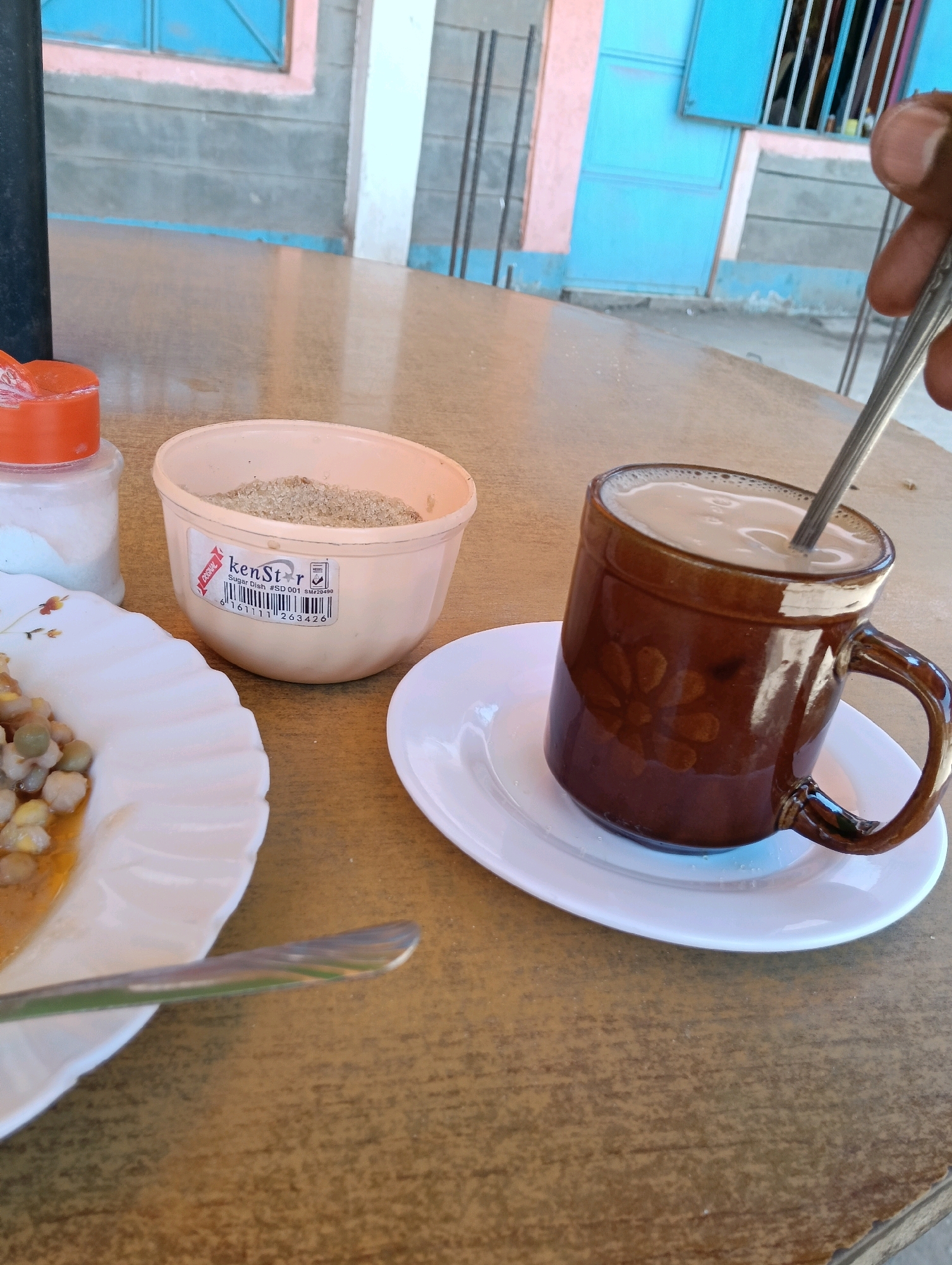 A close-up, eye-level shot captures a casual outdoor dining scene on a wooden table in Zombe/Mwitika ward, Kenya. The primary focus is a steaming mug of what appears to be milky tea or coffee, presented in a brown ceramic mug with a floral design, resting on a white saucer. A spoon is partially submerged in the beverage. To the left, a white plate holds a portion of a meal consisting of chickpeas and corn in a light sauce. Adjacent to the plate is a pale pink plastic container filled with light brown sugar, labeled "kenStar Sugar Dish #SD 001 SN220499". A shaker with an orange lid, containing white granular material (likely salt), stands near the sugar dish. In the background, a building with blue and pink accents is visible, along with a glimpse of metal bars and a black cylindrical object, possibly a bottle or part of a structure. The lighting suggests it's daytime.