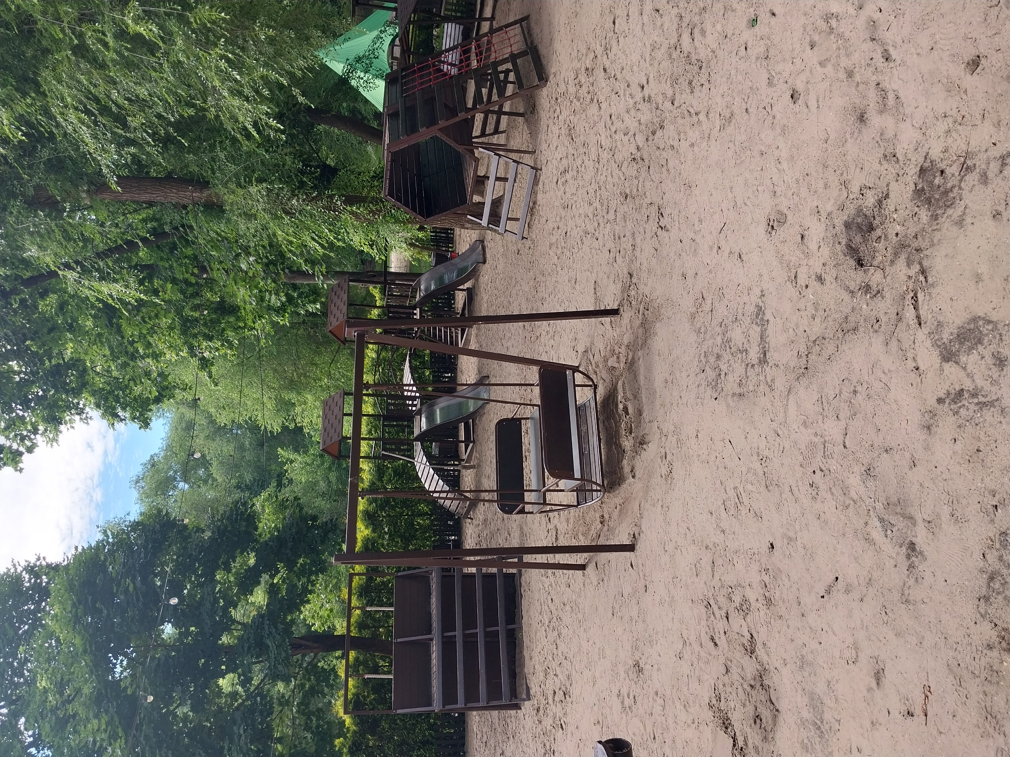 The image shows a children's playground in Dnipro, Ukraine. The playground has a sandy surface and is equipped with various play structures, including a swing set with two seats, a multi-level climbing structure with a slide, and a tiered seating area. The structures are made of wood and metal, with a brown and green color scheme. The playground is surrounded by trees, indicating an outdoor location, possibly in a park or residential area. The lighting suggests it is daytime, with natural sunlight filtering through the foliage. There are no people visible in the image, but footprints in the sand suggest recent use of the playground. The overall scene depicts a well-maintained, yet quiet, recreational space.