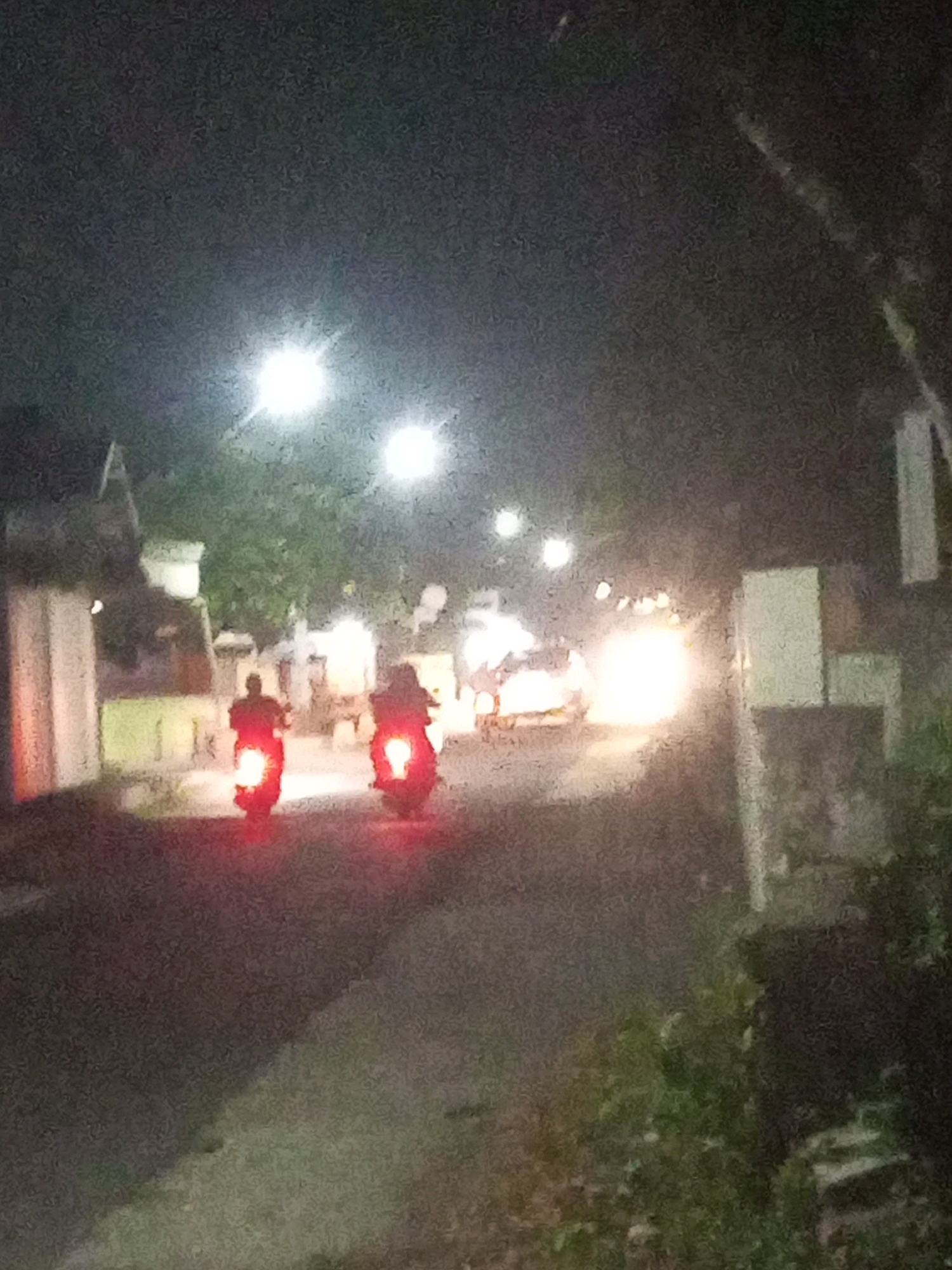 This is a low-light, slightly blurry photograph taken at night on a street in Ngasem, Indonesia. Two figures on motorbikes are visible, their red taillights illuminating the asphalt road. The motorbike on the left appears to be closer to the camera than the one on the right. Buildings with white walls and some greenery line the sides of the street. Several bright lights, possibly streetlights or headlights, are scattered throughout the scene, creating glare and further obscuring details. The overall impression is of a quiet, ordinary street scene at night.